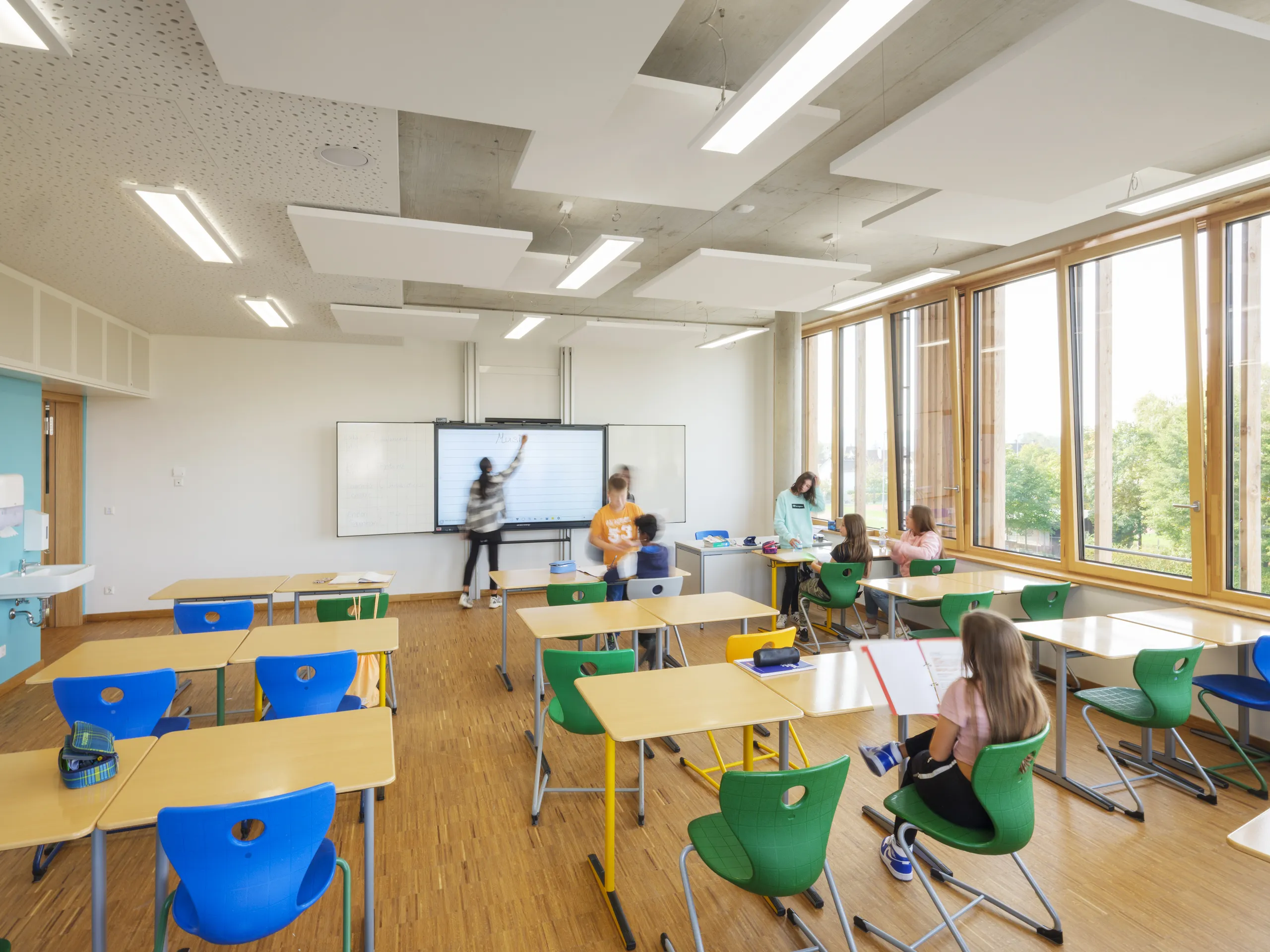 A contemporary classroom featuring white rectangular acoustic ceiling rafts hung beneath an exposed concrete soffit, with students working at desks near tall timber-framed glazing