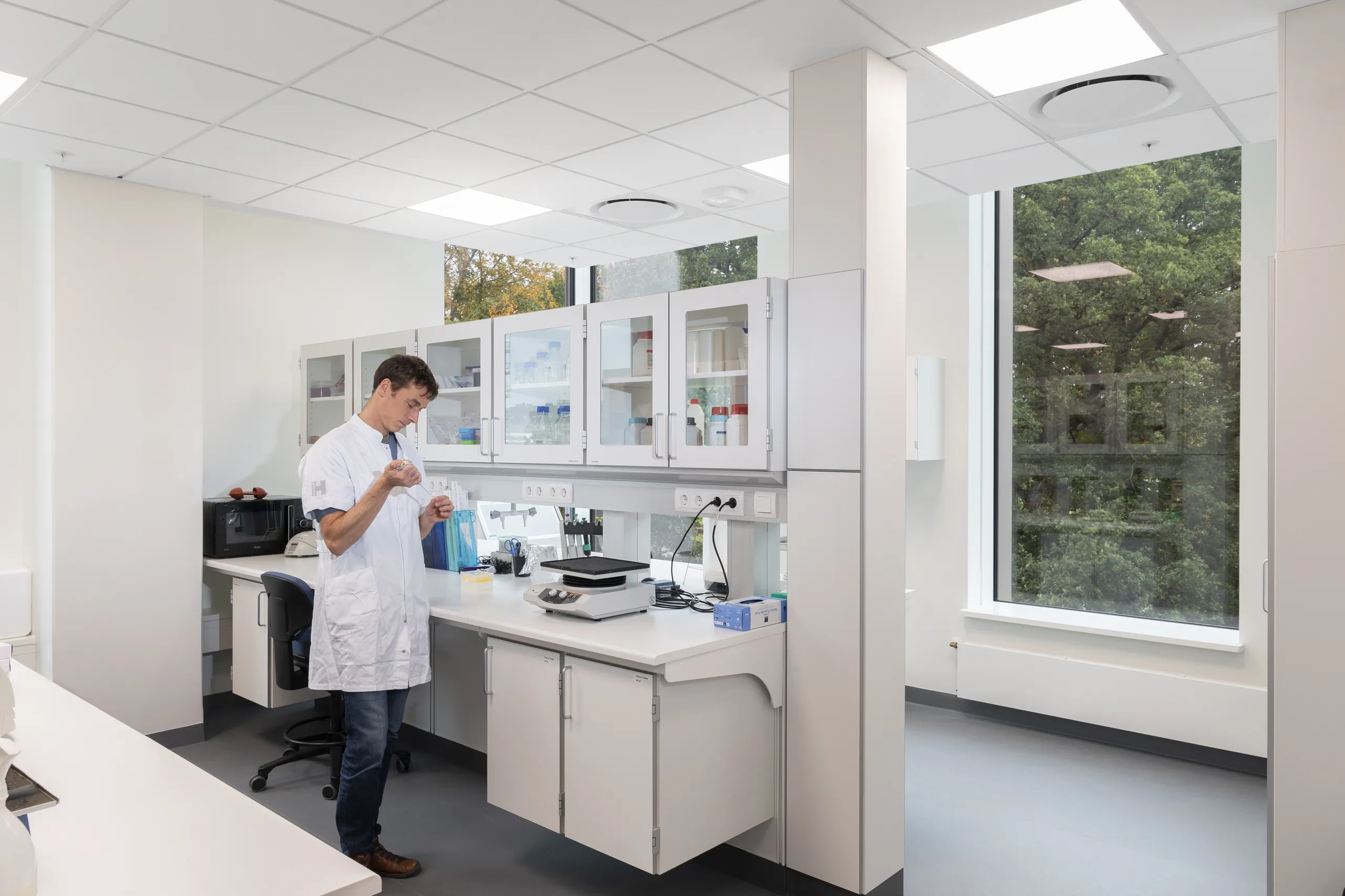 A clean, bright laboratory with a white suspended acoustic ceiling and large windows overlooking greenery. A researcher in a lab coat works at a bench surrounded by cabinets, equipment, and integrated services, supported by a quiet, controlled acoustic environment.