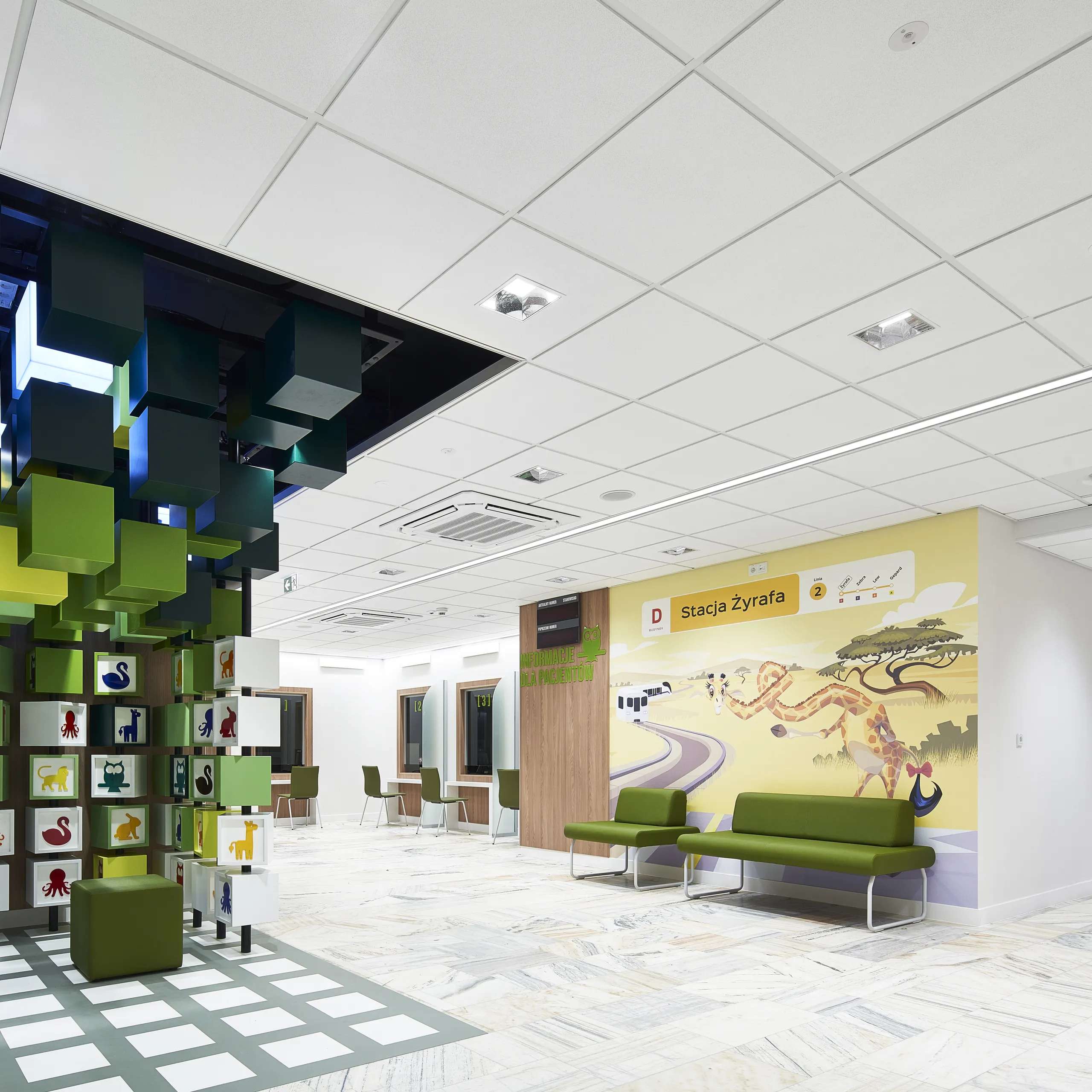 A bright paediatric waiting area featuring a suspended installation of colourful green cubes, playful animal graphics on wall panels, and a clean white acoustic ceiling with integrated lighting.