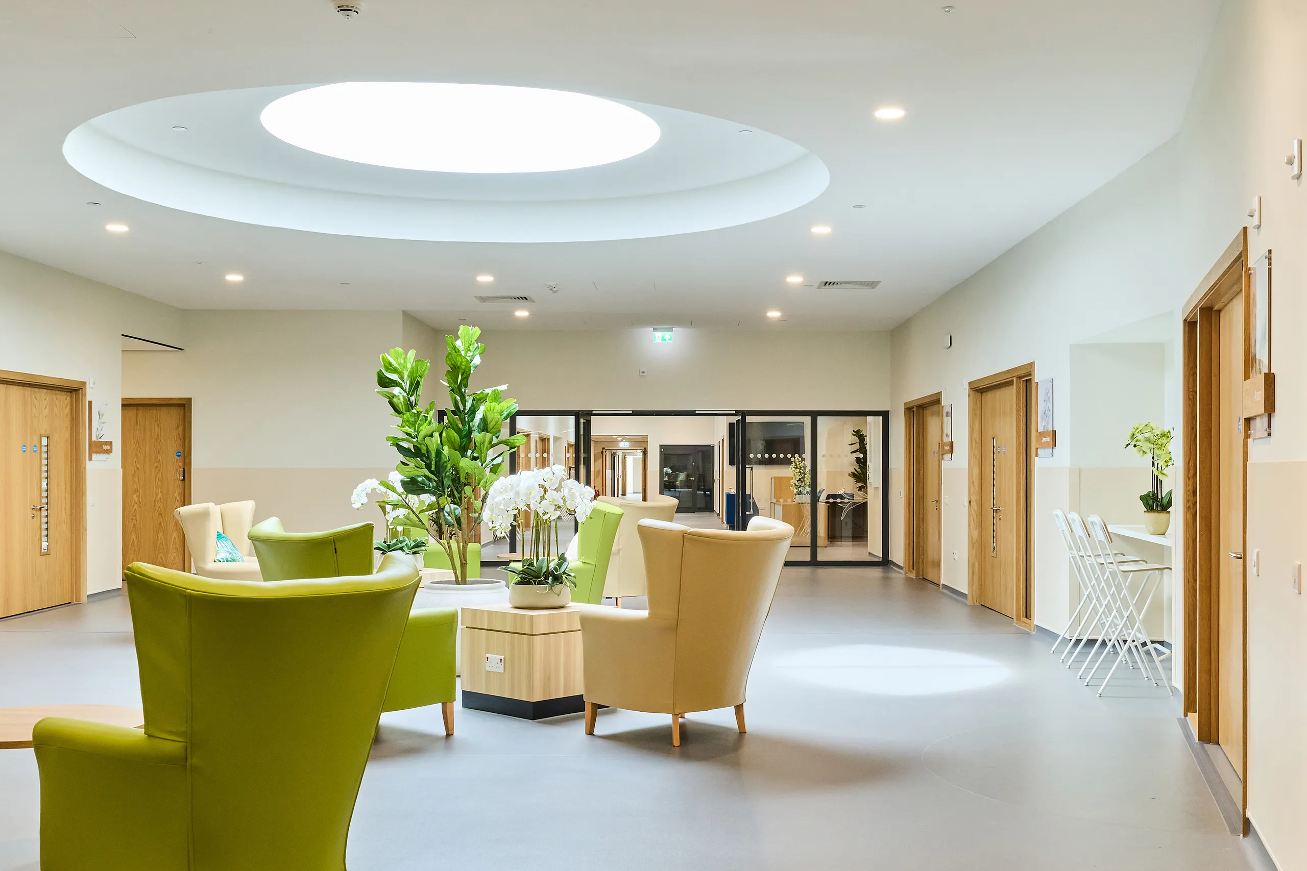A bright healthcare lounge with soft seating arranged beneath a large circular skylight, featuring a smooth acoustic ceiling that supports a calm, low-noise environment. Light wood doors, indoor plants, and neutral finishes create a welcoming, restorative space.