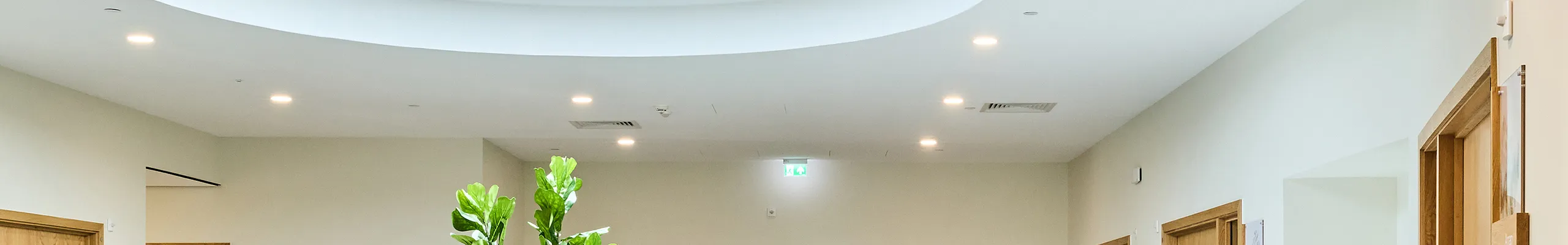 A bright healthcare lounge with soft seating arranged beneath a large circular skylight, featuring a smooth acoustic ceiling that supports a calm, low-noise environment. Light wood doors, indoor plants, and neutral finishes create a welcoming, restorative space.