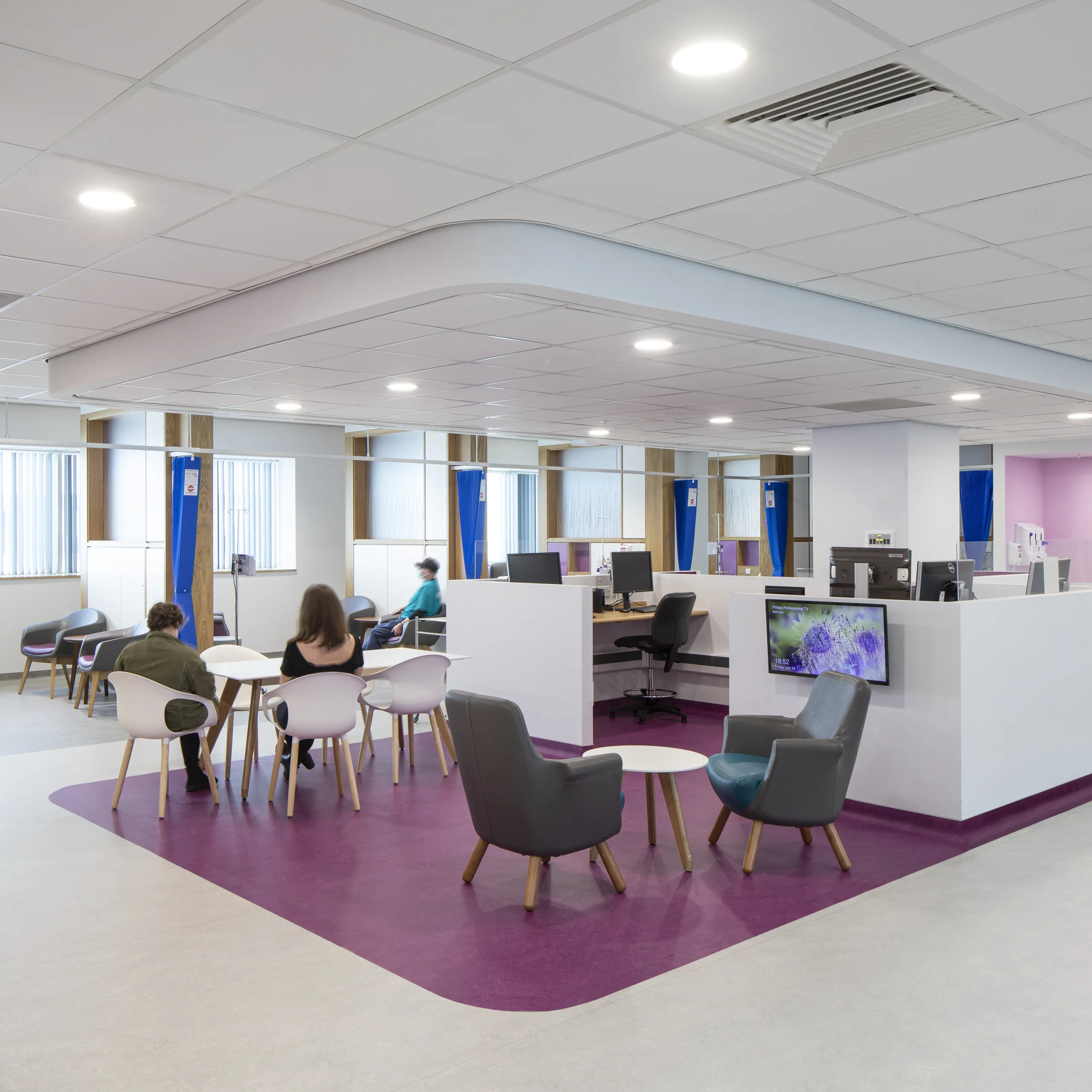 A modern hospital waiting area featuring a suspended white acoustic ceiling with integrated lighting, purple zoning on the floor, workstations, and a distinctive green acoustic ceiling island above the central desks.