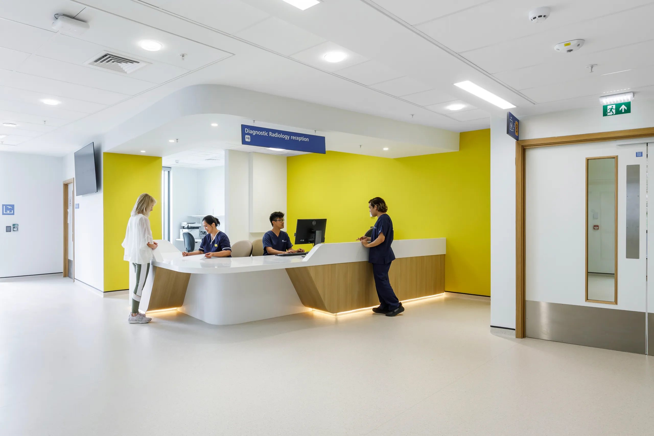 Diagnostic radiology reception area with nurses assisting patients, yellow feature wall and white acoustic ceiling panels.