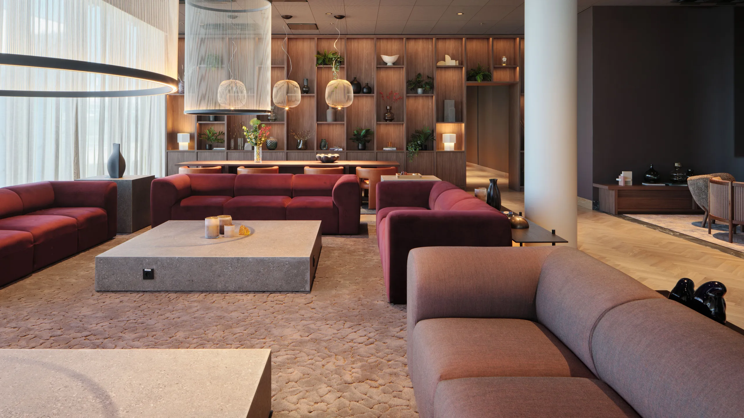 Hotel lounge with modular acoustic ceiling tiles, red sofas, pendant lighting, and a wooden shelving wall with plants and decorative objects.