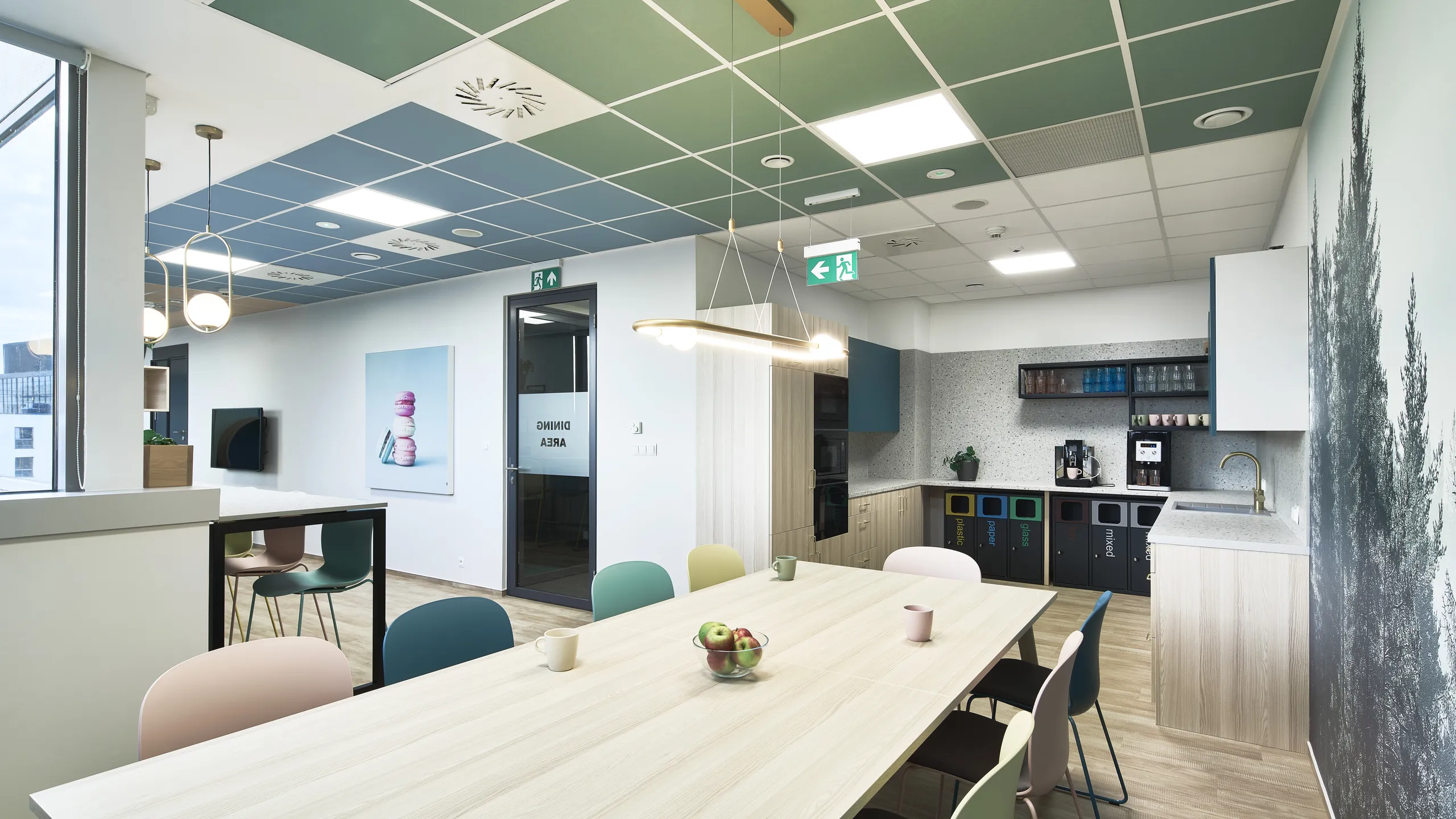Modern office kitchen with a long wooden table, colorful chairs, and a kitchenette. Green and blue ceiling panels enhance the space.