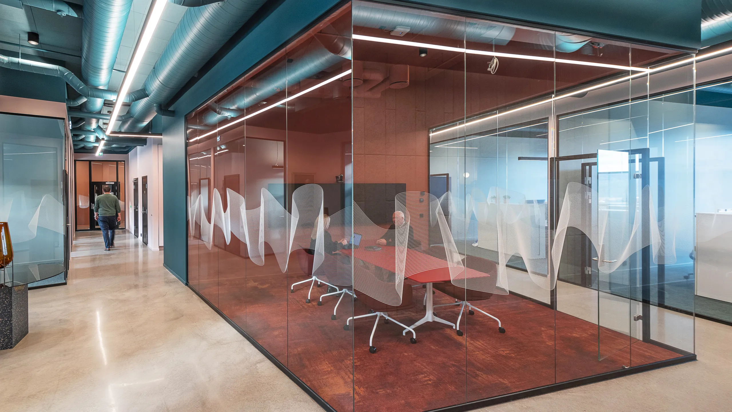 Office corridor with exposed services and acoustic ceiling elements, glass meeting rooms with coloured finishes, and a collaborative workspace visible through transparent partitions.