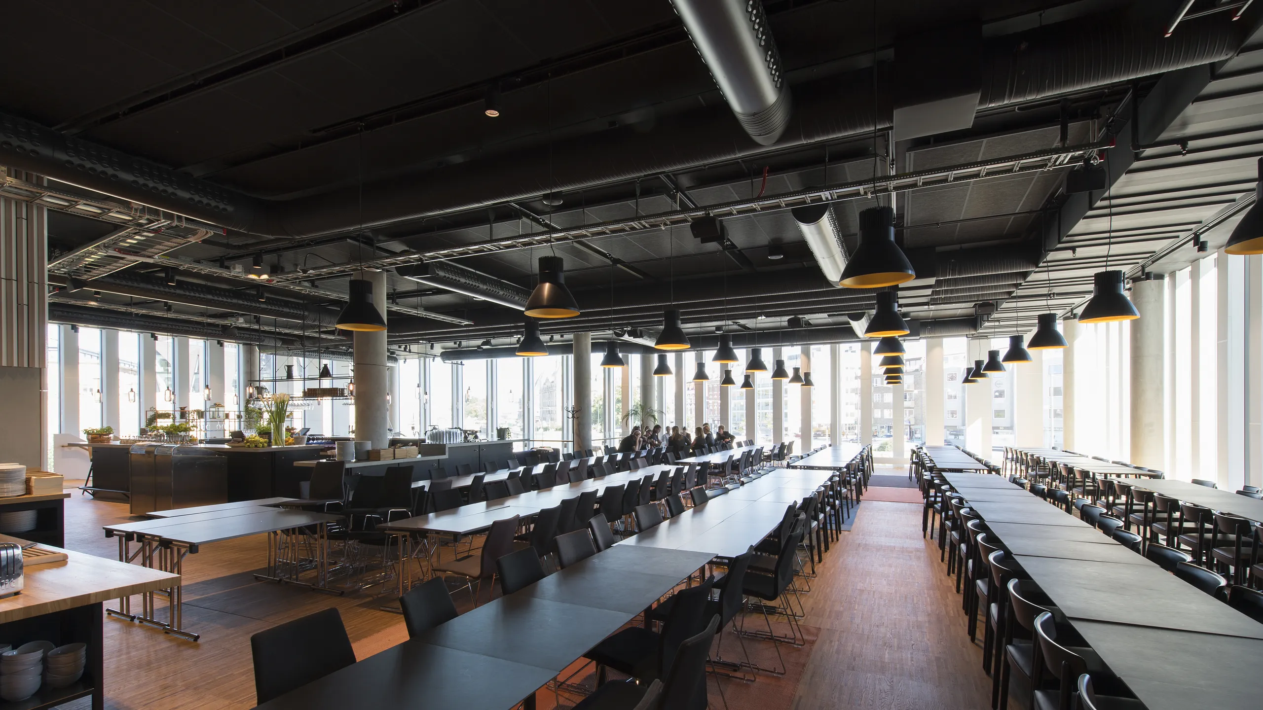 Spacious, modern cafeteria with a black suspended ceiling, black tables and chairs, large windows, and overhead lighting, featuring a clean, industrial design.