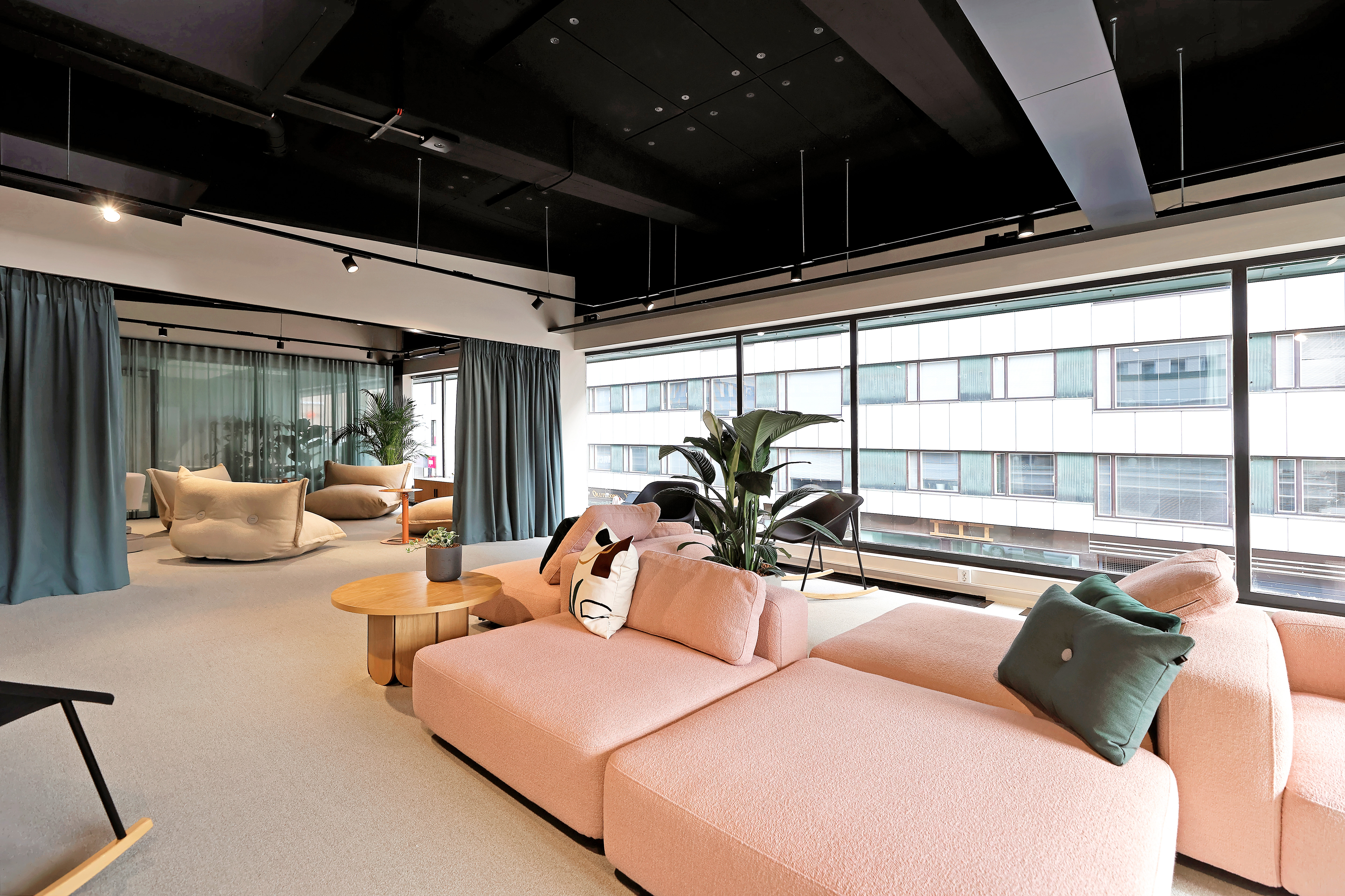 Modern lounge with black suspended ceiling, pink sofas, green cushions, and large windows. Potted plants add greenery; soft lighting creates a cozy ambiance.