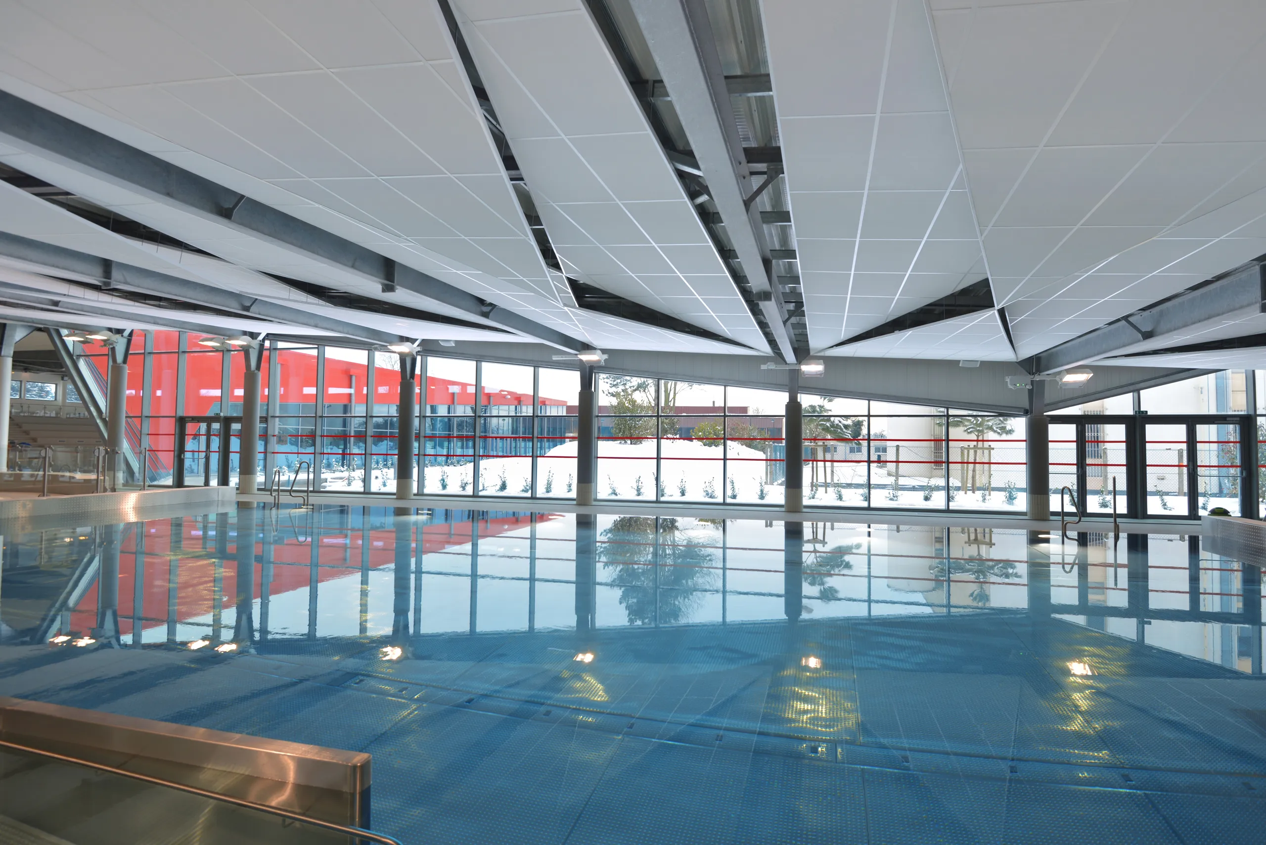 Indoor swimming pool with large windows and exposed steel structure, featuring suspended white acoustic ceiling panels reflected in the calm water surface.