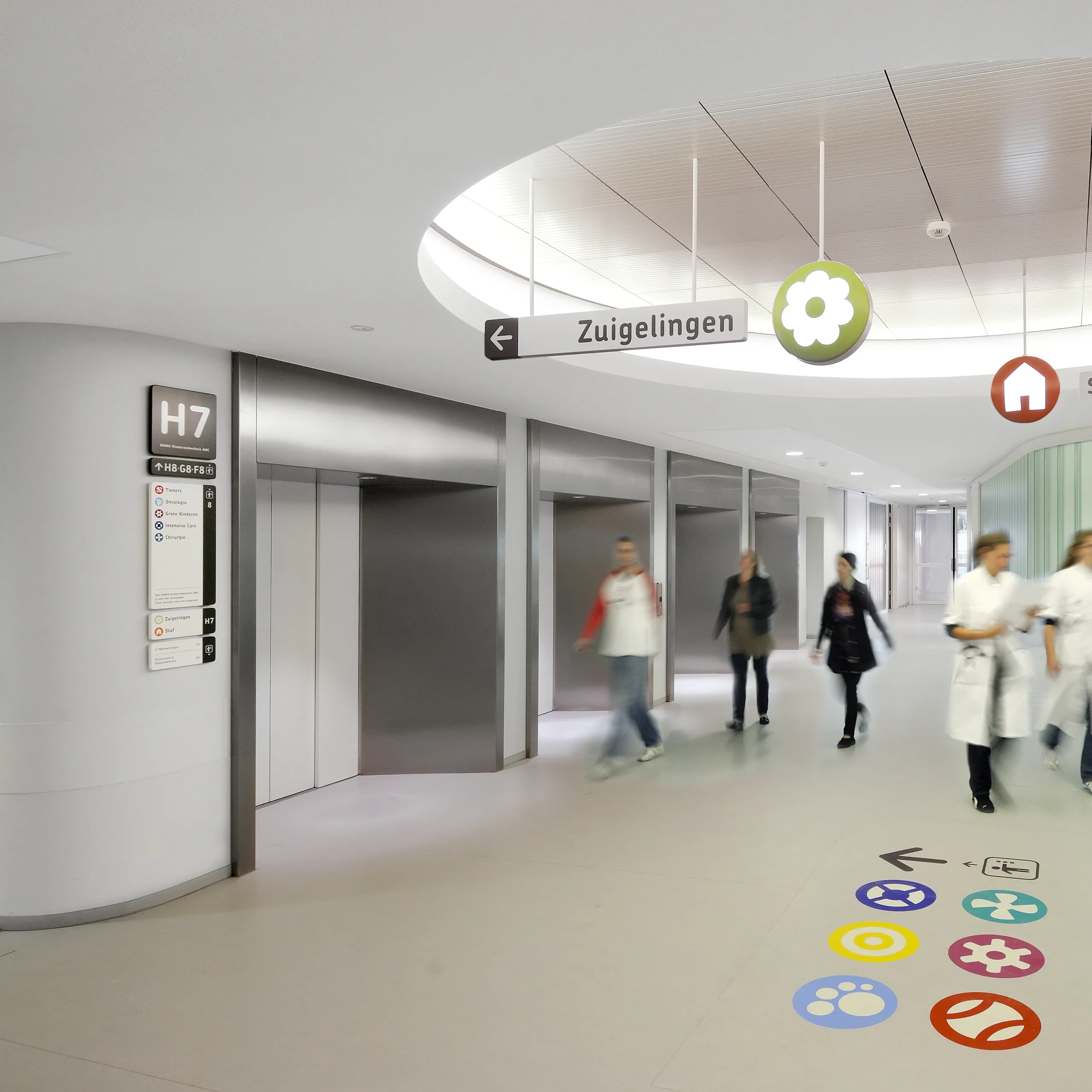 Hospital corridor with multiple elevators, seamless monolithic acoustic ceiling feature, wayfinding signage, and medical staff walking through.
