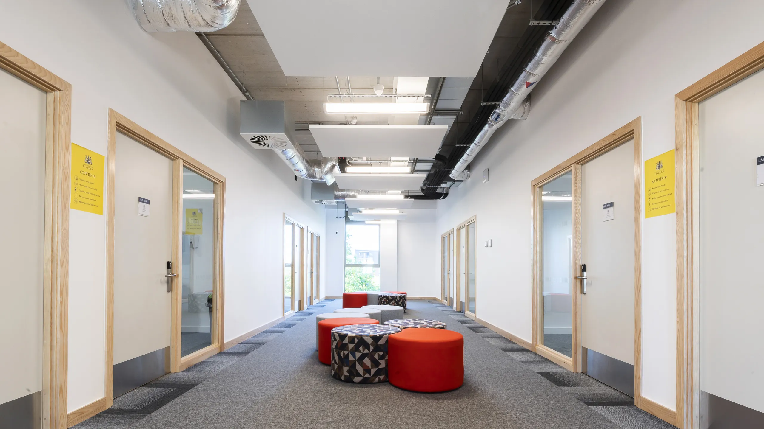 A modern hallway with square floating ceiling panels, exposed piping, wooden door frames, colorful ottomans, and carpet flooring leading to a bright window.