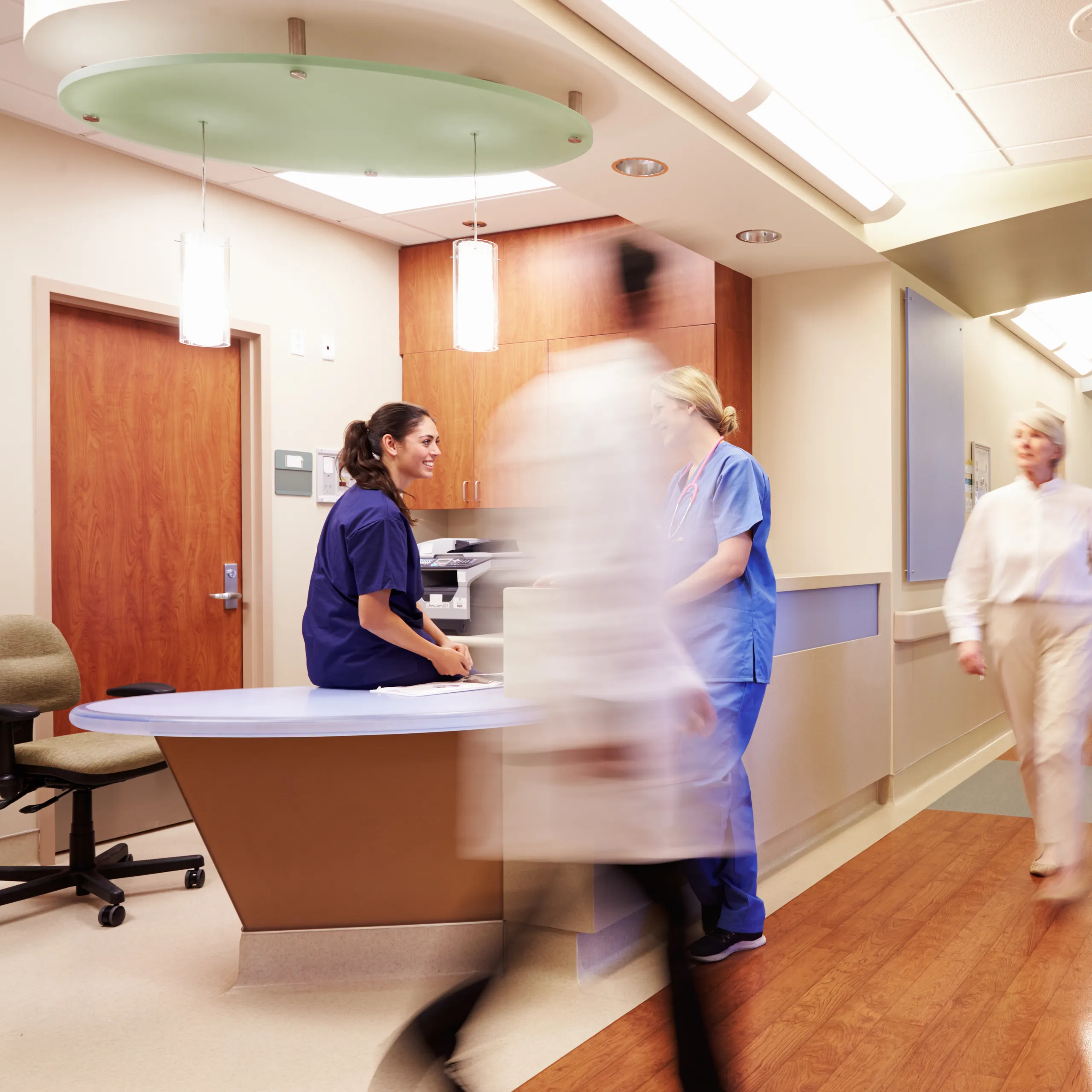 Hospital corridor with nurses and doctors walking past a central reception desk, featuring a suspended circular acoustic panel above.