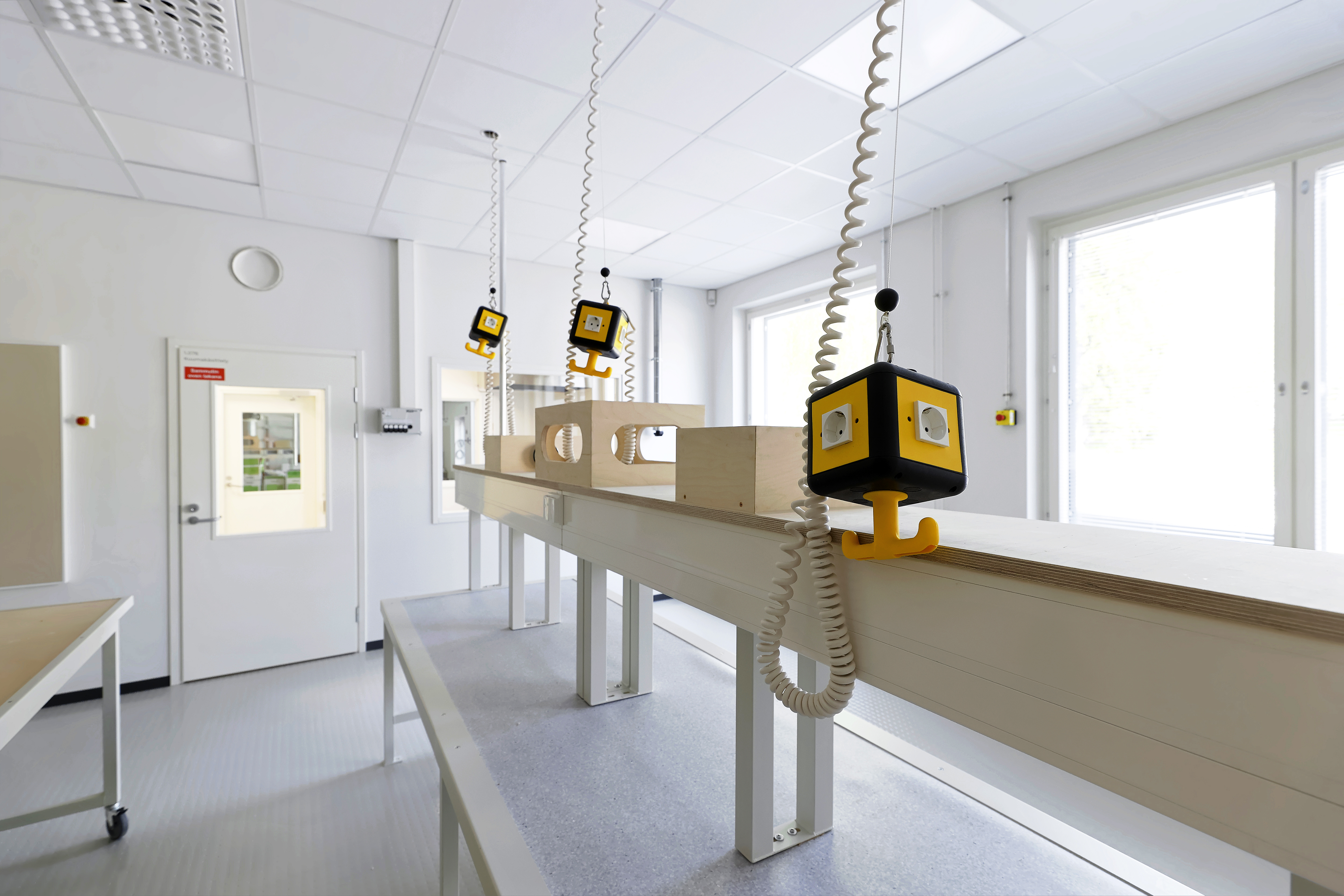 Workshop or technical classroom with white acoustic ceiling tiles, workbenches with suspended power units, and large windows providing natural light.