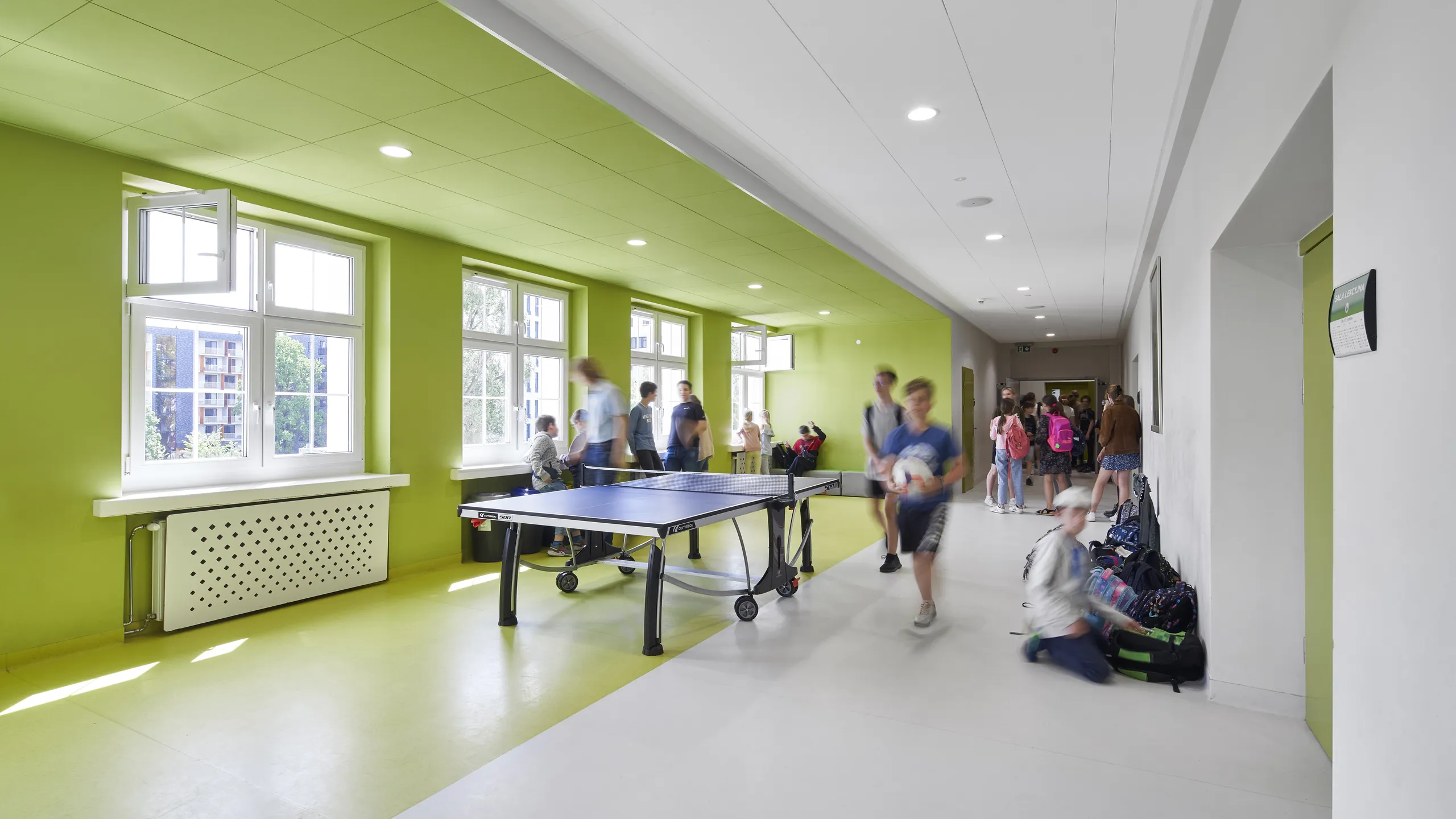 A bright hallway with bright green ceiling and walls, featuring a table tennis setup. Two blurred figures walk by, suggesting motion and activity.