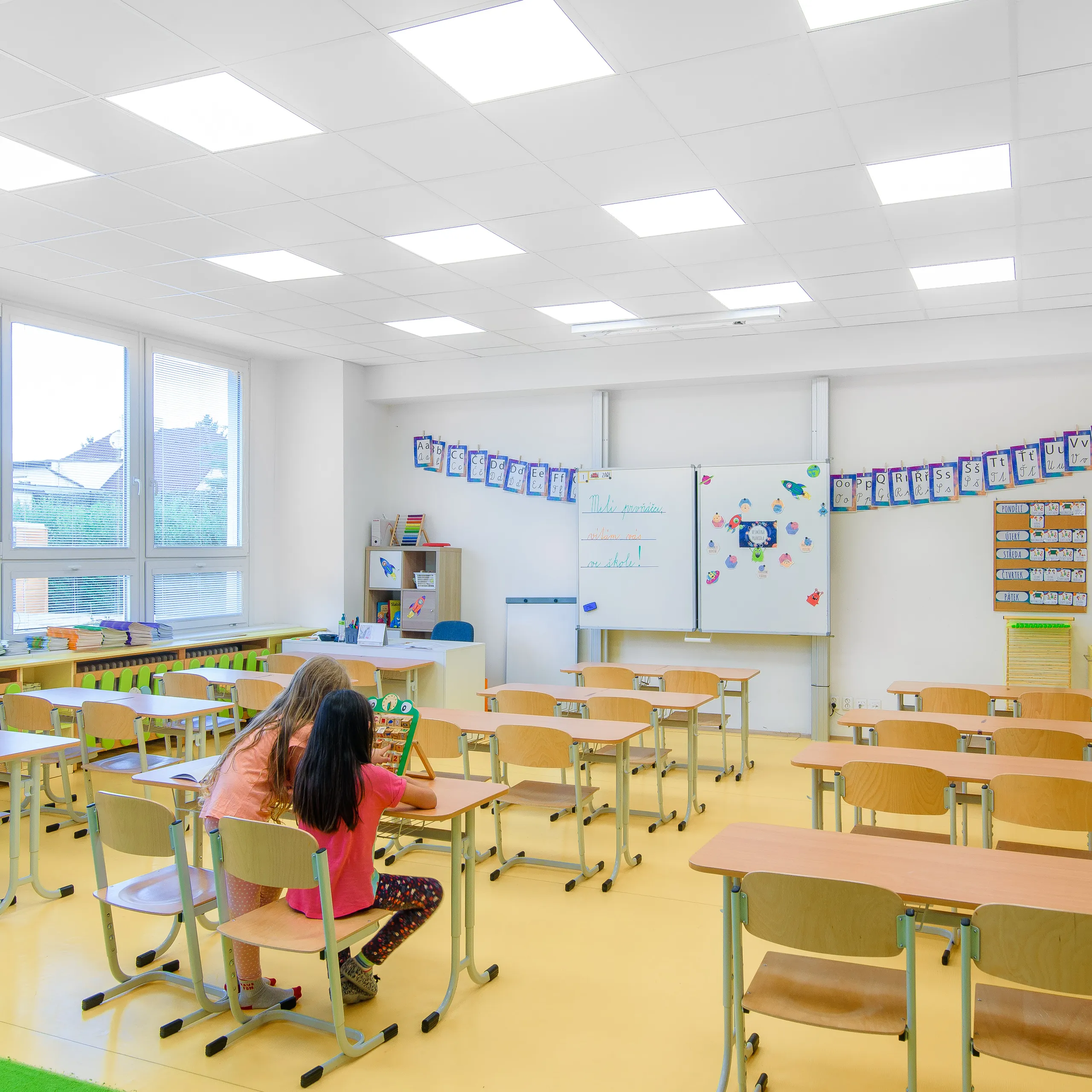 A colourful primary classroom featuring a white acoustic ceiling tile grid and bright yellow flooring, with two children working together at the front and large windows bringing in natural light.