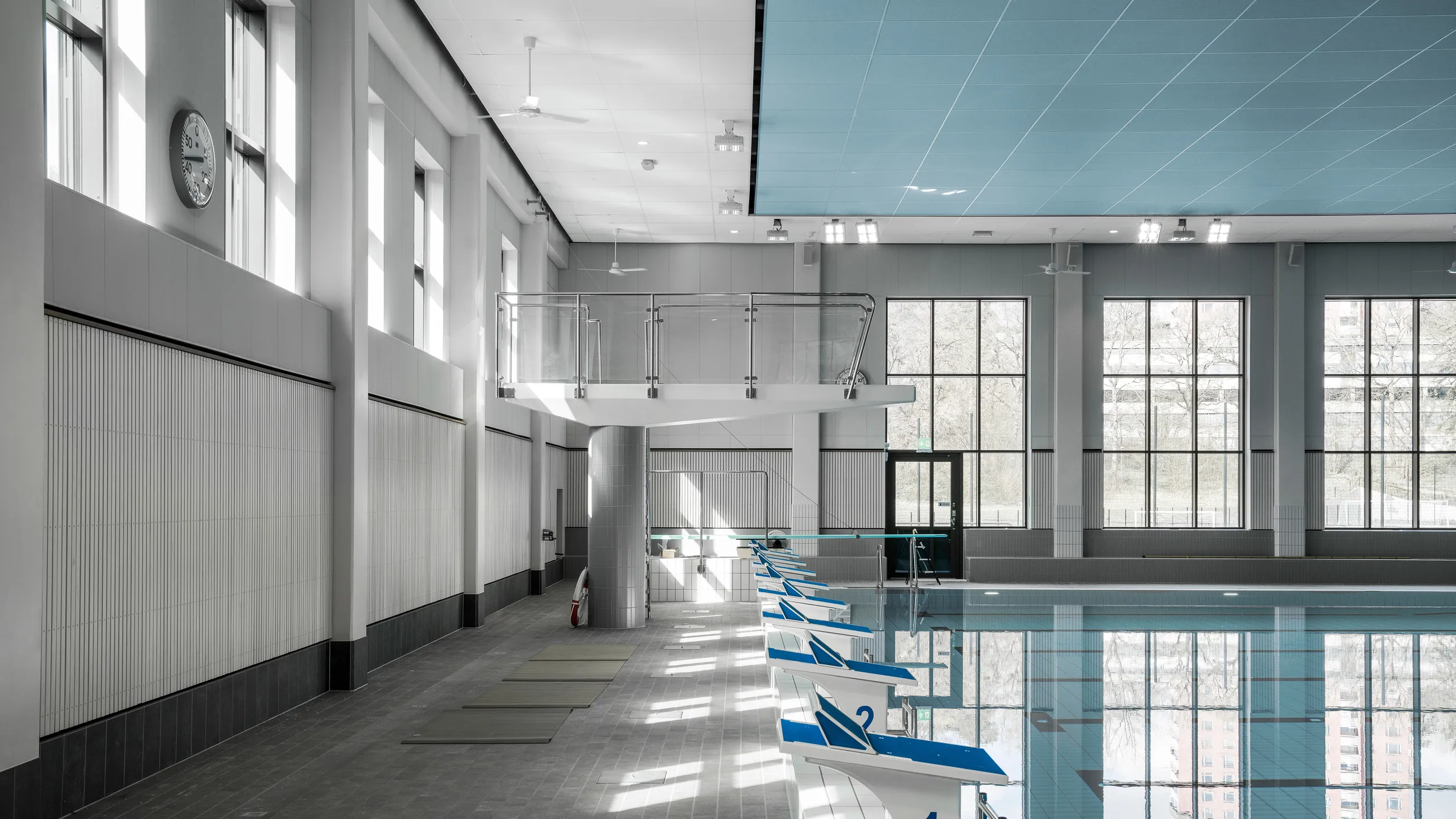 Indoor swimming pool with a blue acoustic ceiling, diving platform, lane markings, and starting blocks aligned along the pool edge in a bright, daylit hall.