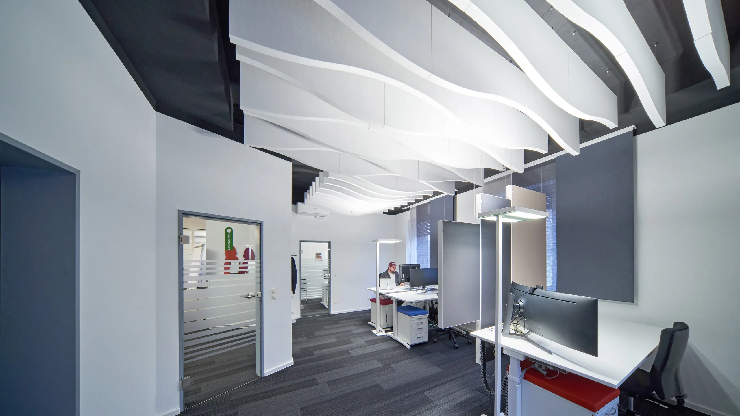Office workspace with desks and white curved waved acoustic ceiling baffles suspended above the interior.