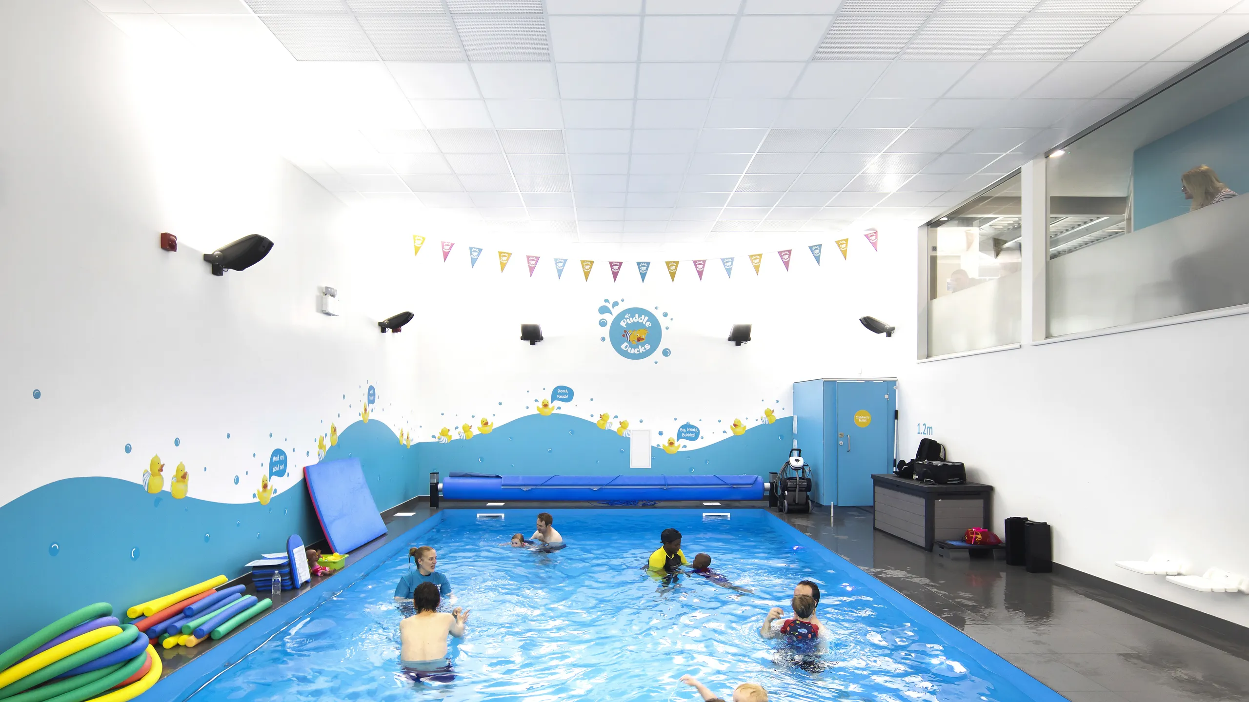 Indoor swimming pool with children and instructor, colorful wall decorations, and pool toys around. Bright overhead lighting and glass observation area.