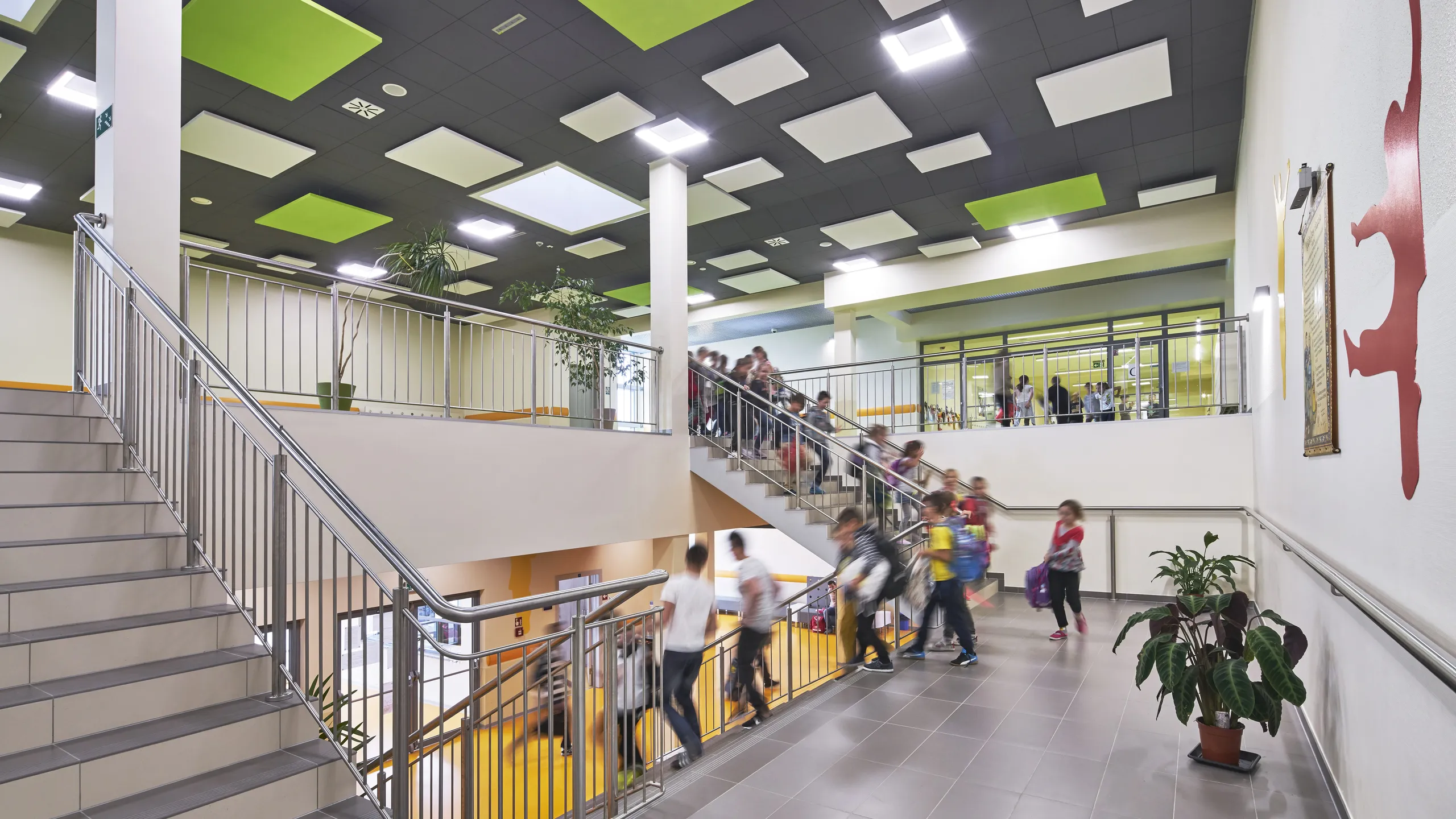 A modern school interior with students moving on a stairway. The ceiling features green and white panels, and there's a plant near the stairs.