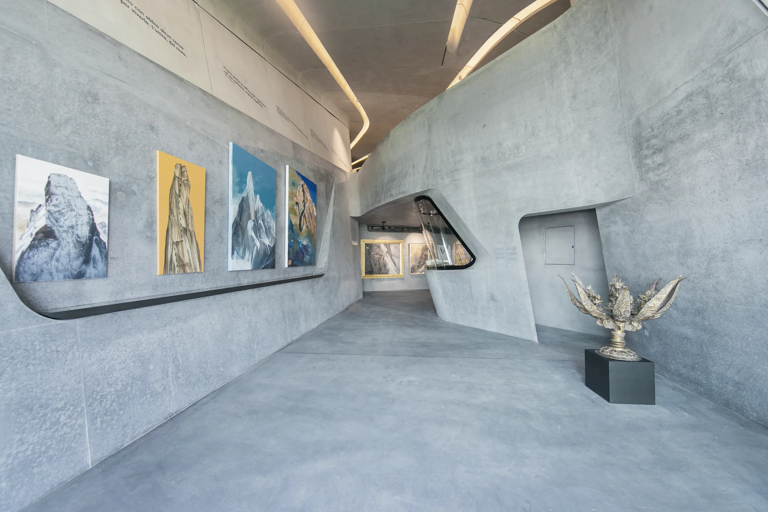 Modern museum gallery with sculpted concrete walls, a curved circulation path, and artworks and sculptures displayed along the walls under recessed linear lighting.