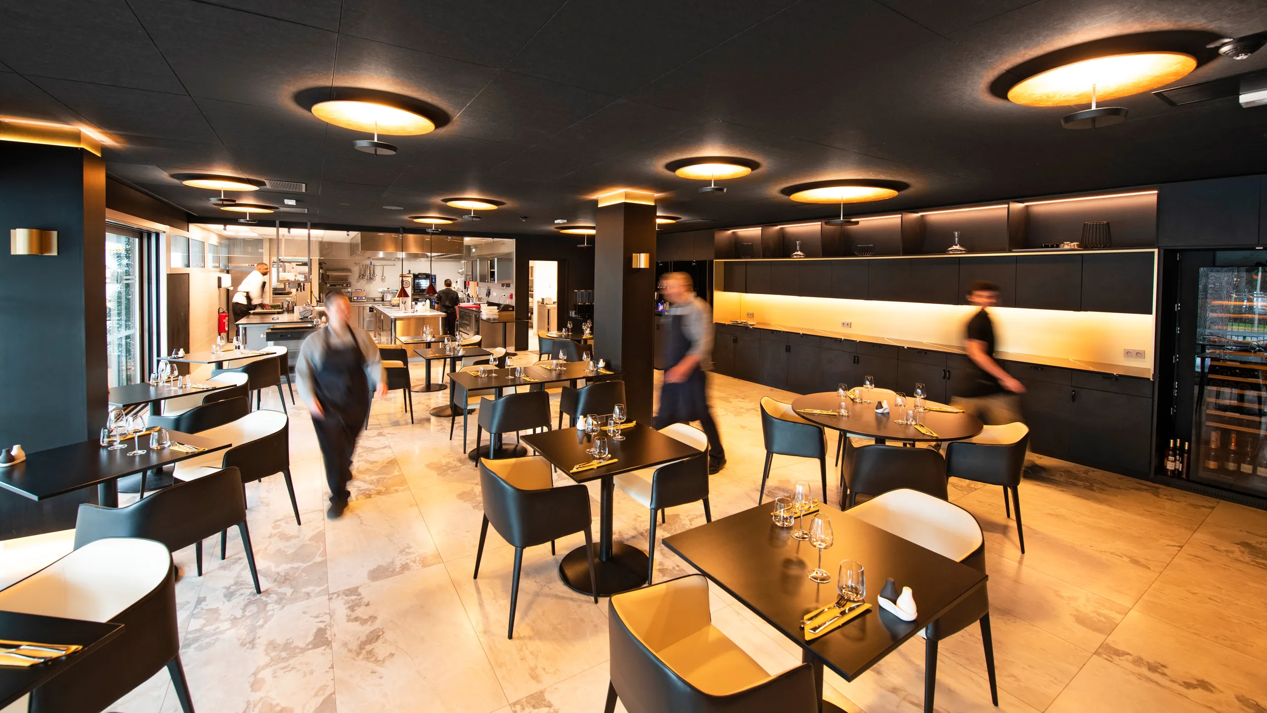 Modern restaurant interior with black suspended ceiling, several tables and chairs, dim lighting, warm tones, and blurred figures of staff walking.