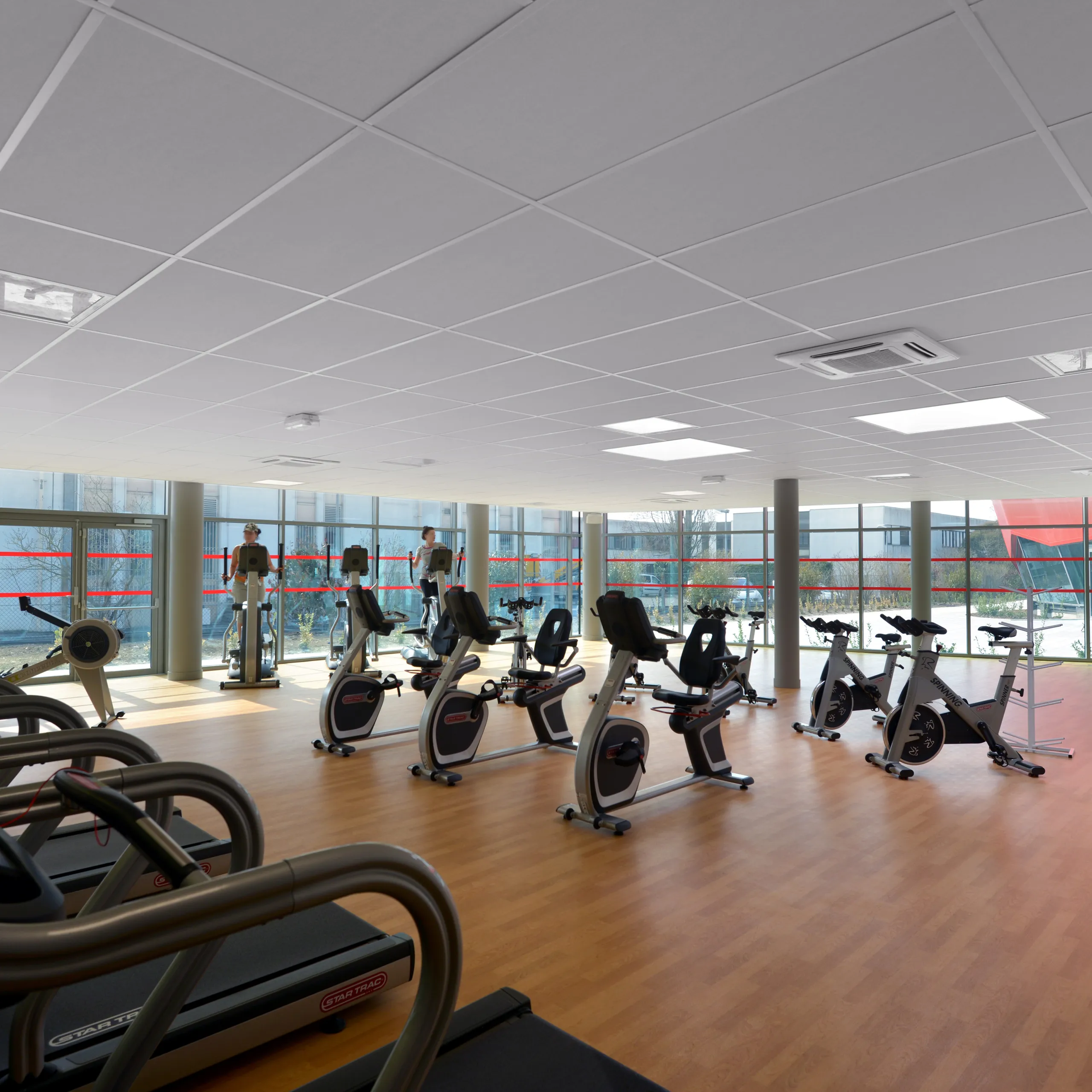 Fitness studio with treadmills and stationary bikes arranged on a wooden floor beneath a white acoustic tile ceiling and large glazed façade overlooking the outdoors.