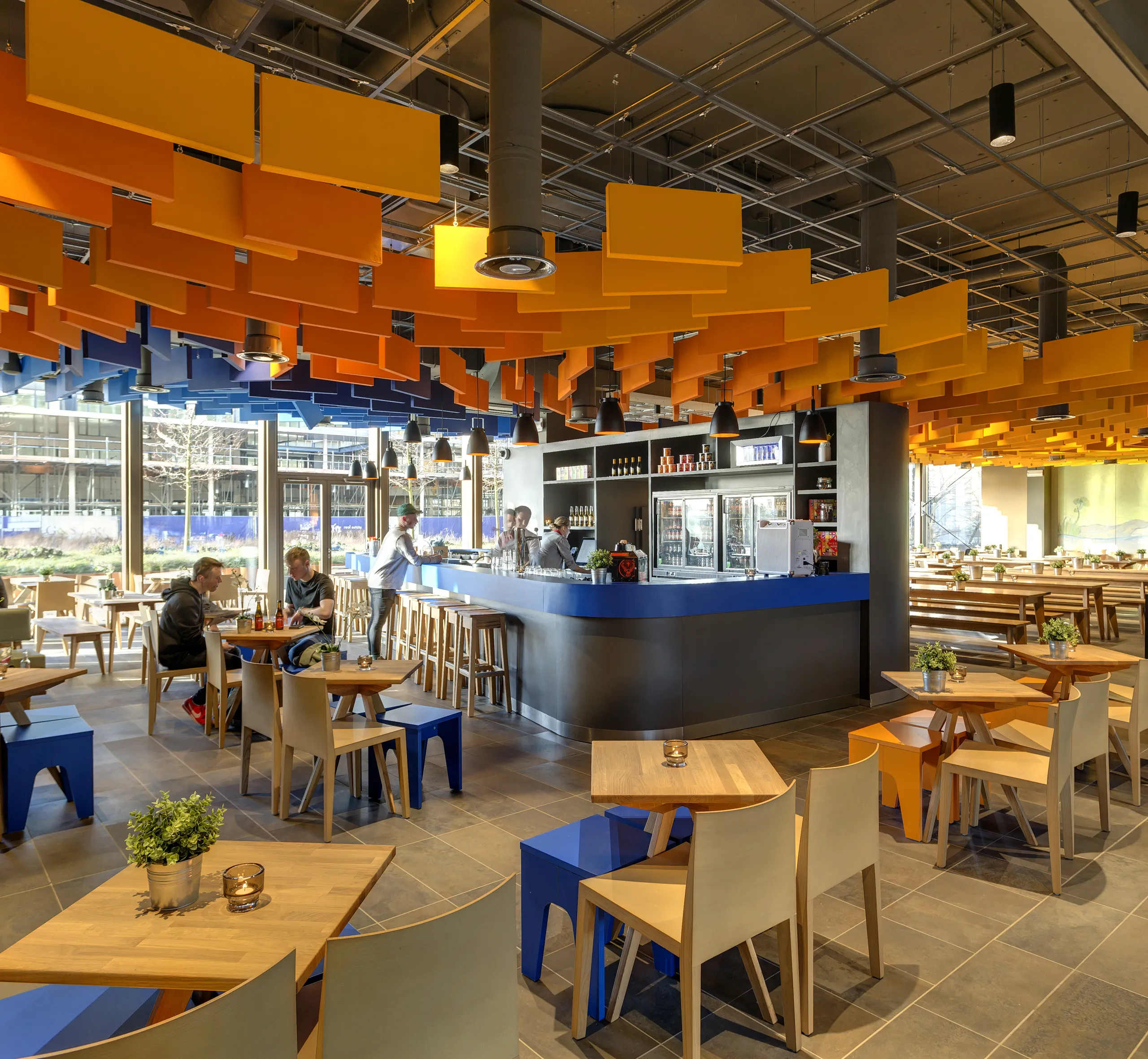 Restaurant interior with suspended orange and blue acoustic baffles forming a dynamic ceiling, central bar area, and seating arranged throughout a bright, open dining space.