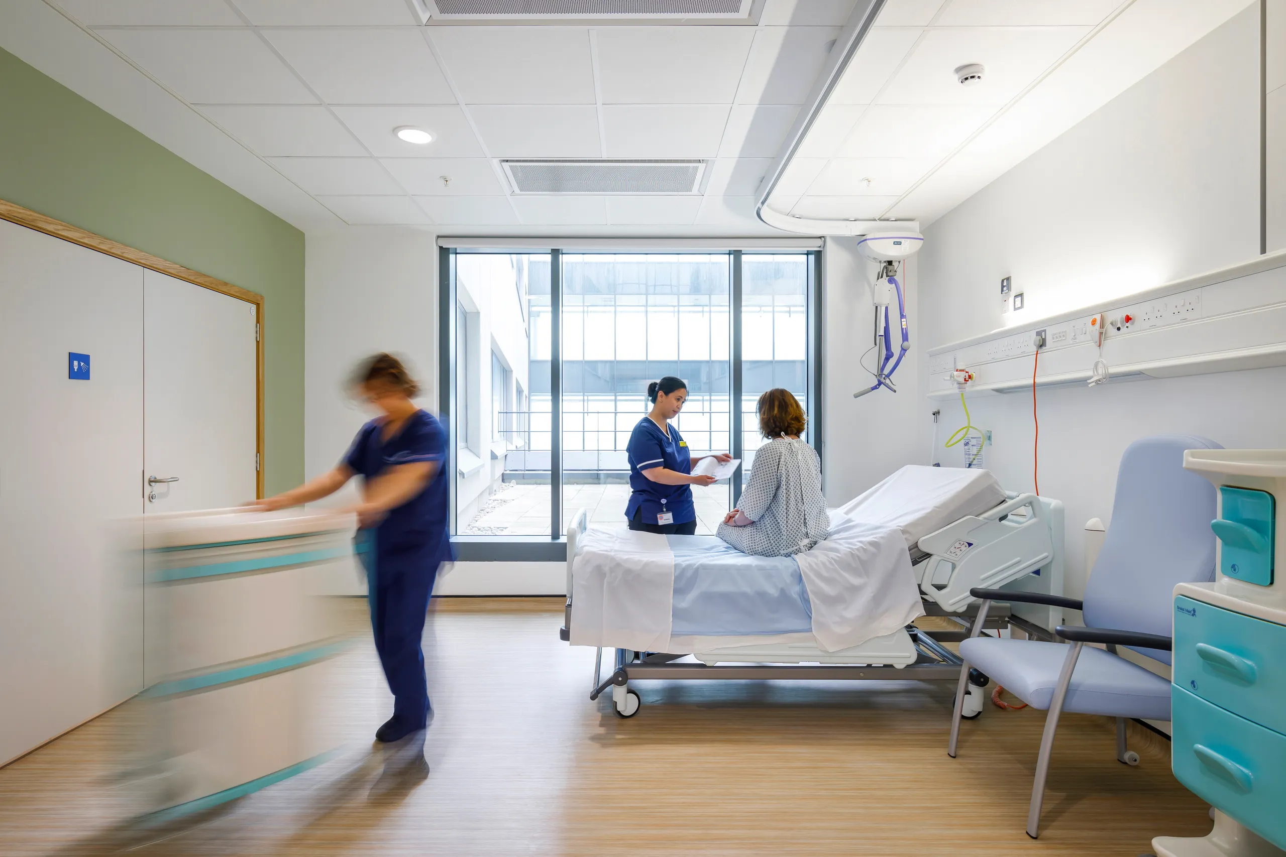 A bright hospital patient room with a suspended acoustic ceiling grid, where a nurse consults a patient seated on the bed while another staff member moves equipment. Large windows bring in natural light, and medical services line the wall to support a quiet, comfortable care environment.