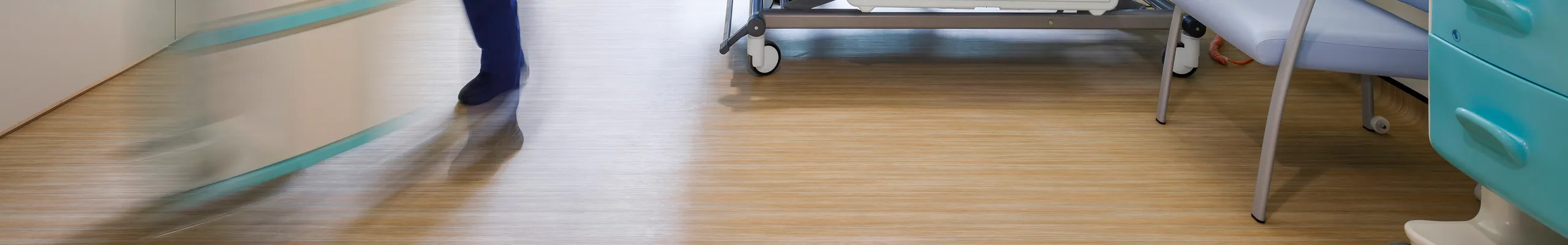 A bright hospital patient room with a suspended acoustic ceiling grid, where a nurse consults a patient seated on the bed while another staff member moves equipment. Large windows bring in natural light, and medical services line the wall to support a quiet, comfortable care environment.