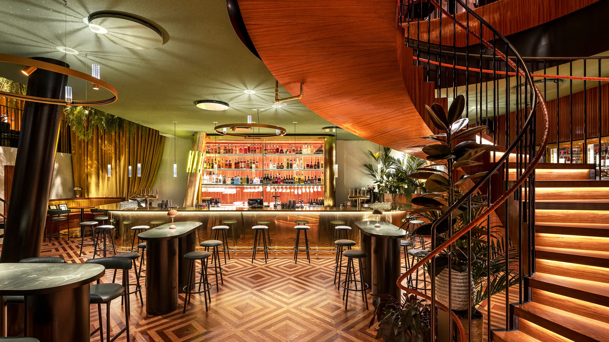 A stylish bar with patterned wooden floors, a spiral staircase, high tables, stools, and a well-stocked, illuminated bar counter in the background.