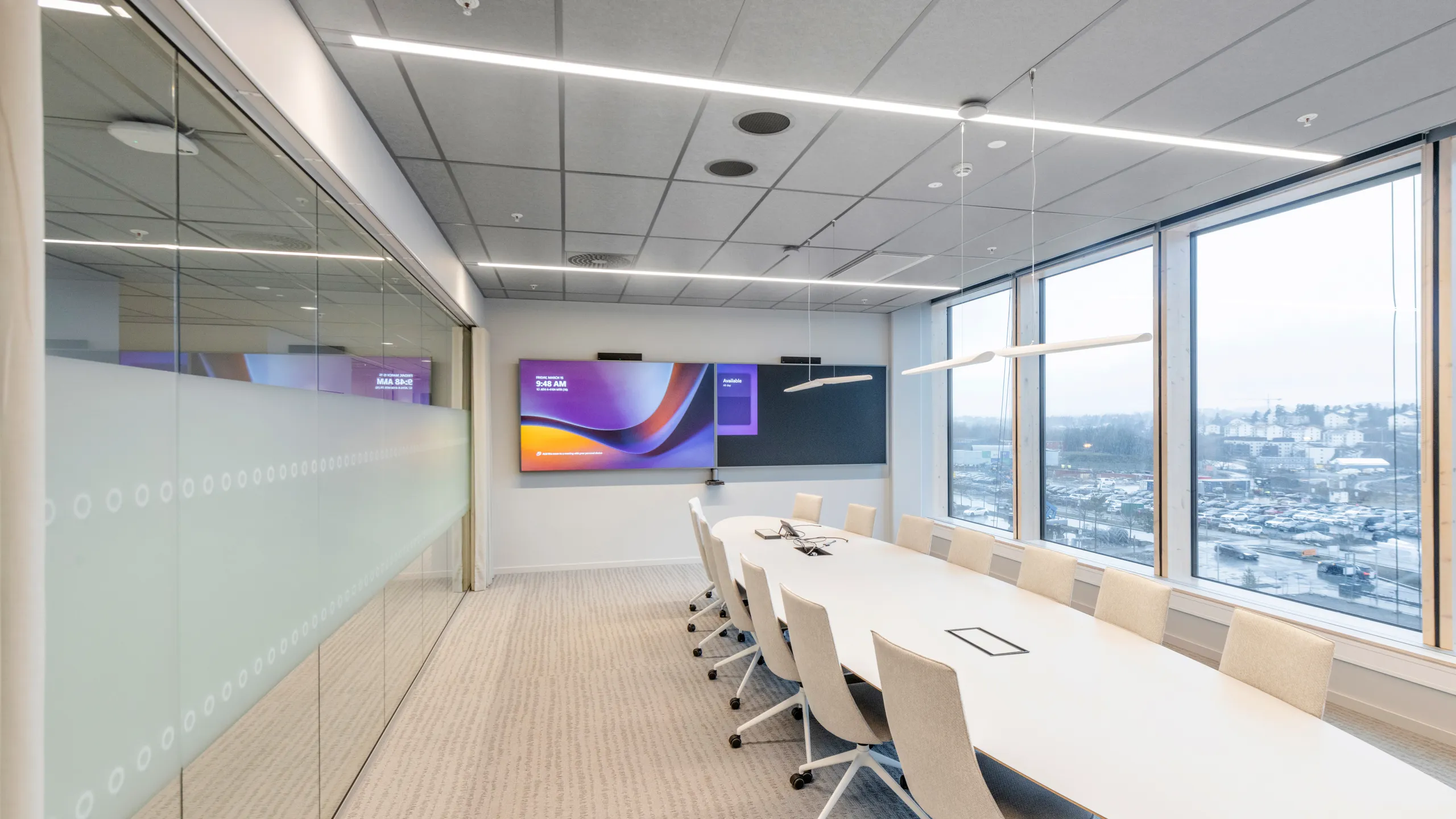 Modern conference room with a long meeting table, large digital display, and a grey acoustic tile ceiling with integrated linear lighting above the space.
