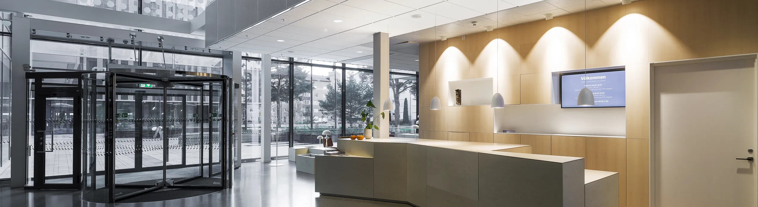 Bright reception area with a suspended acoustic ceiling grid, large glass entrance, wood-panelled front desk, and modern pendant lighting