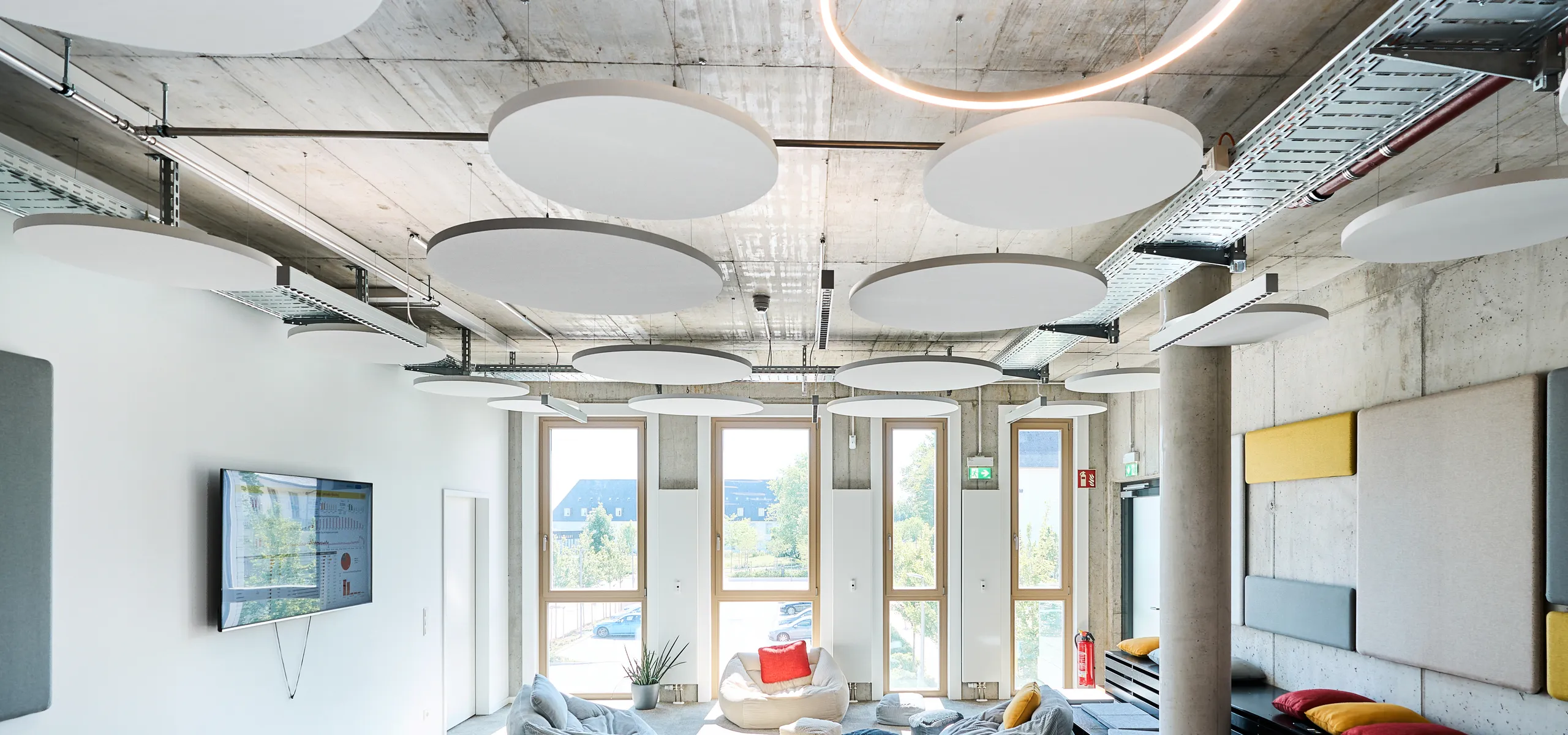 Modern breakout room featuring round acoustic islands on an exposed concrete ceiling, soft seating areas, tiered platforms, and tall windows providing natural lightModern breakout room featuring round acoustic islands on an exposed concrete ceiling, soft seating areas, tiered platforms, and tall windows providing natural light