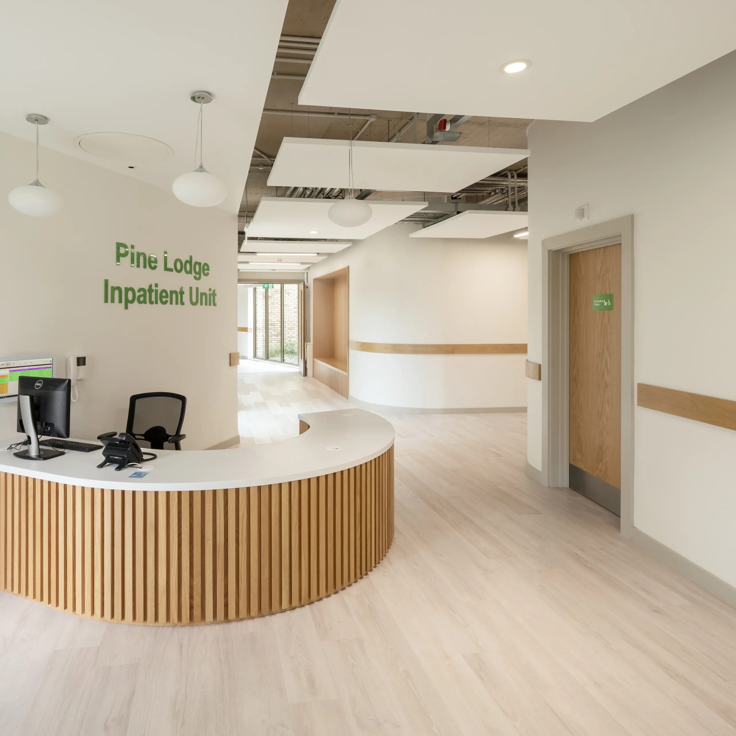 A welcoming inpatient unit reception with a curved wooden desk, pendant lighting and suspended white acoustic ceiling islands.