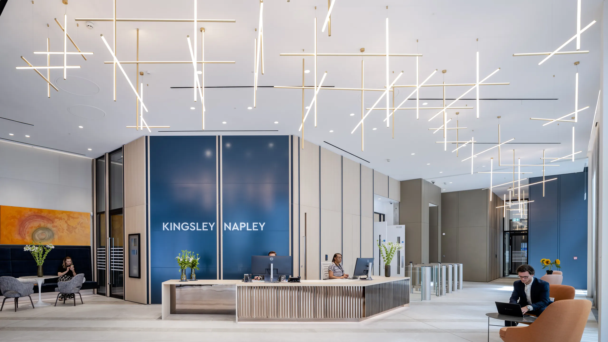 Modern office reception with a seamless white office ceiling, sleek desk, blue panels, seated visitors, and glowing linear ceiling lights.