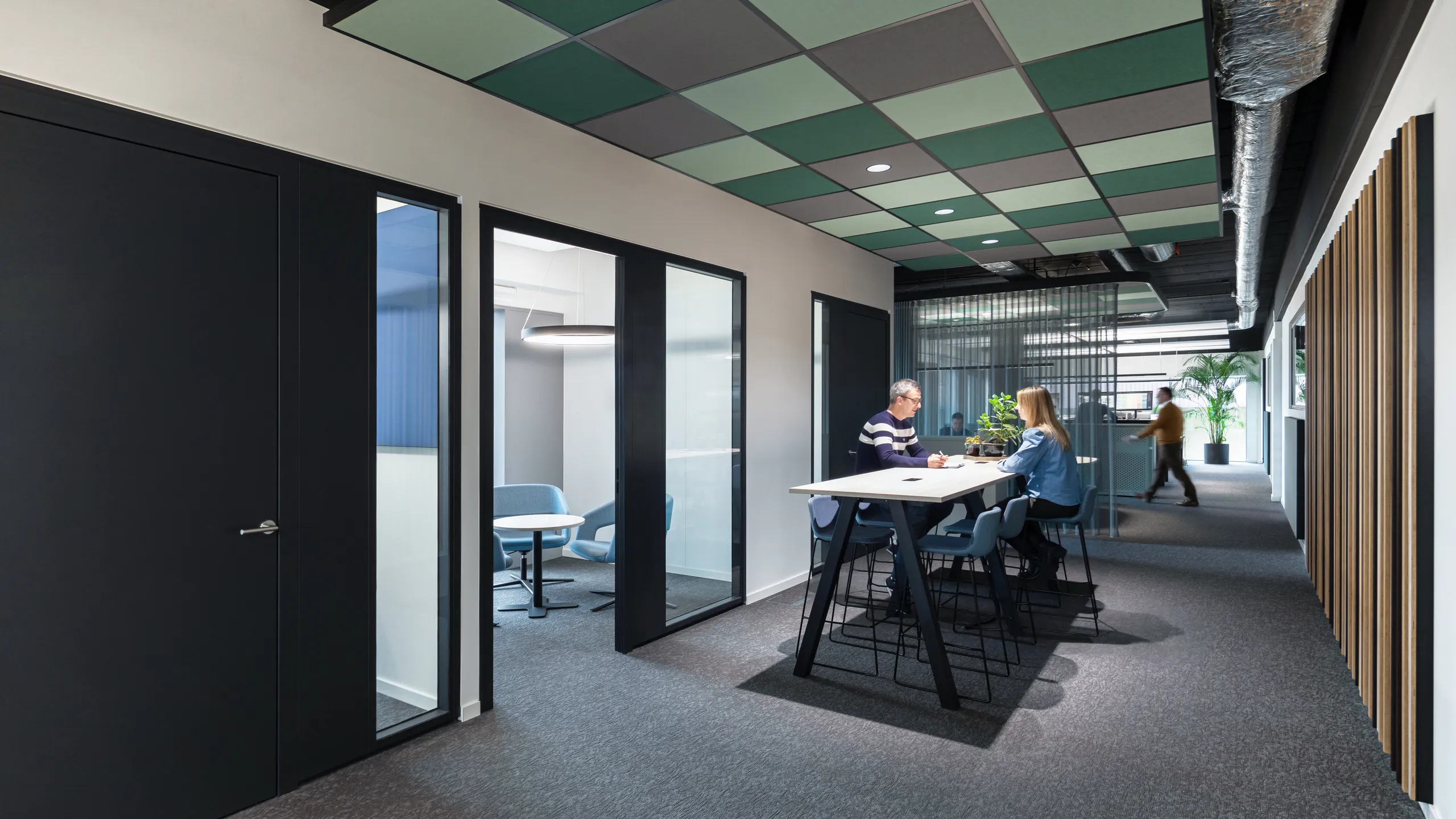 Modern office hallway with black doors, glass-walled meeting rooms, and a high table. Two people are seated, engaged in conversation.