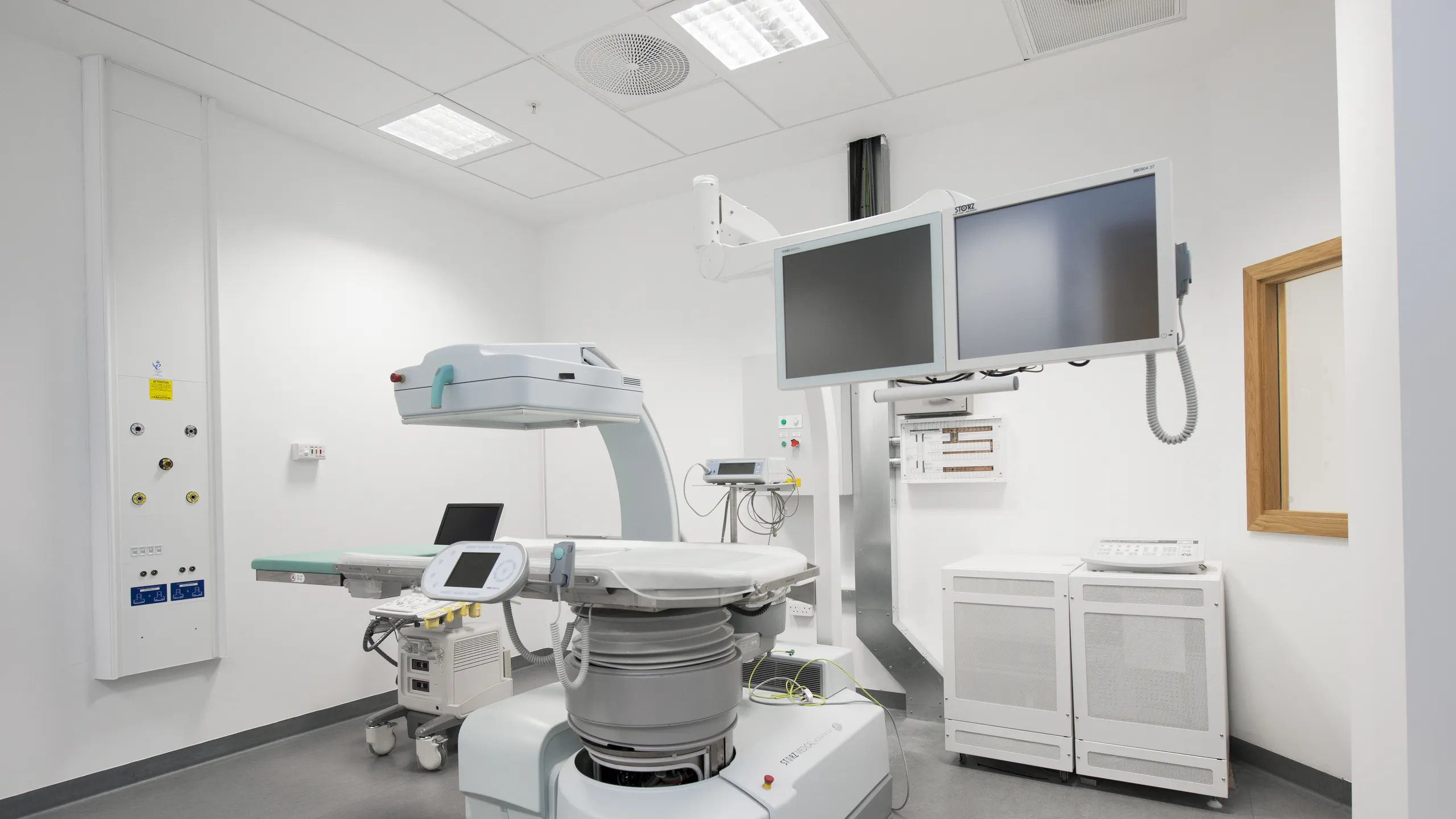 Hospital treatment room with white acoustic ceiling tiles, medical imaging equipment, monitoring screens, and a clean, clinical layout.