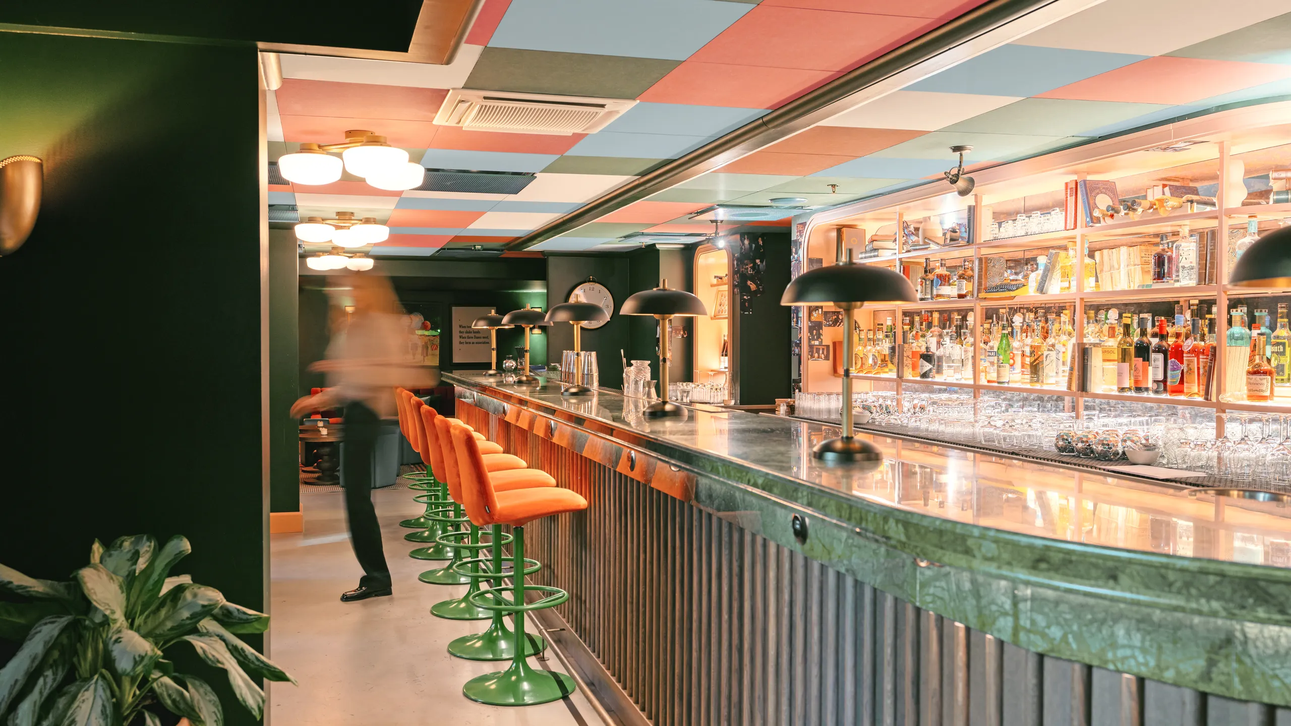 Colourful acoustic ceiling tiles above a stylish bar interior with a long green marble counter, orange bar stools, pendant lamps, and illuminated shelves of bottles behind the bar.