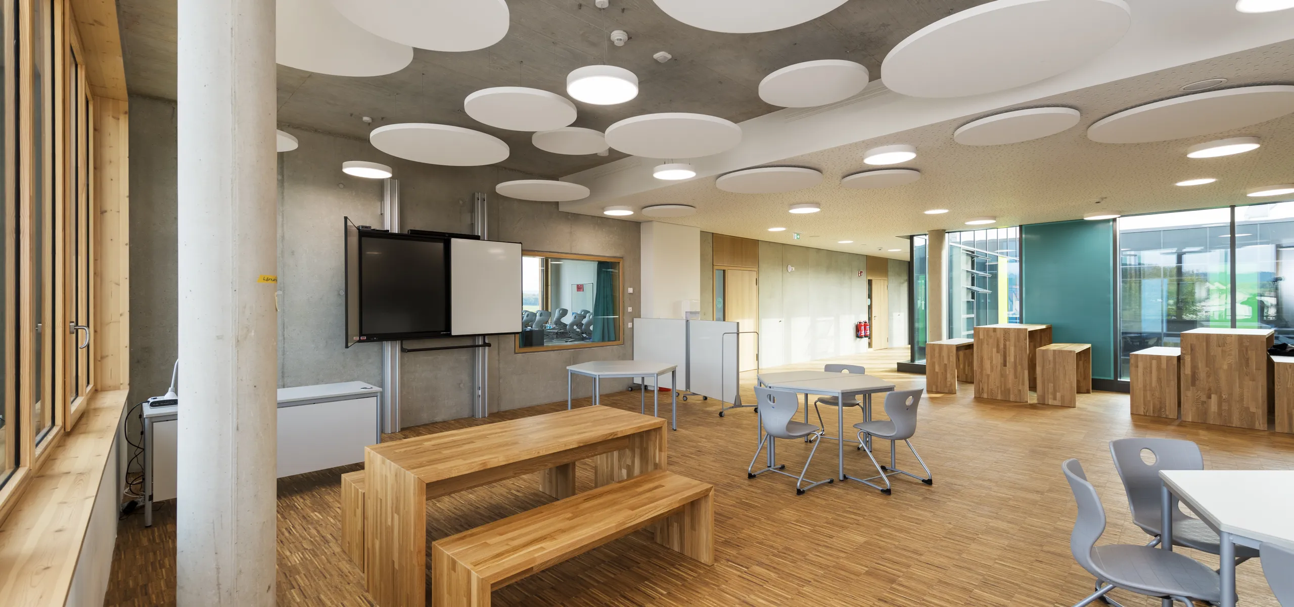 Modern classroom with wooden floors, benches, circular ceiling panels, a large screen, and blue-green walls. Natural light from large windows.