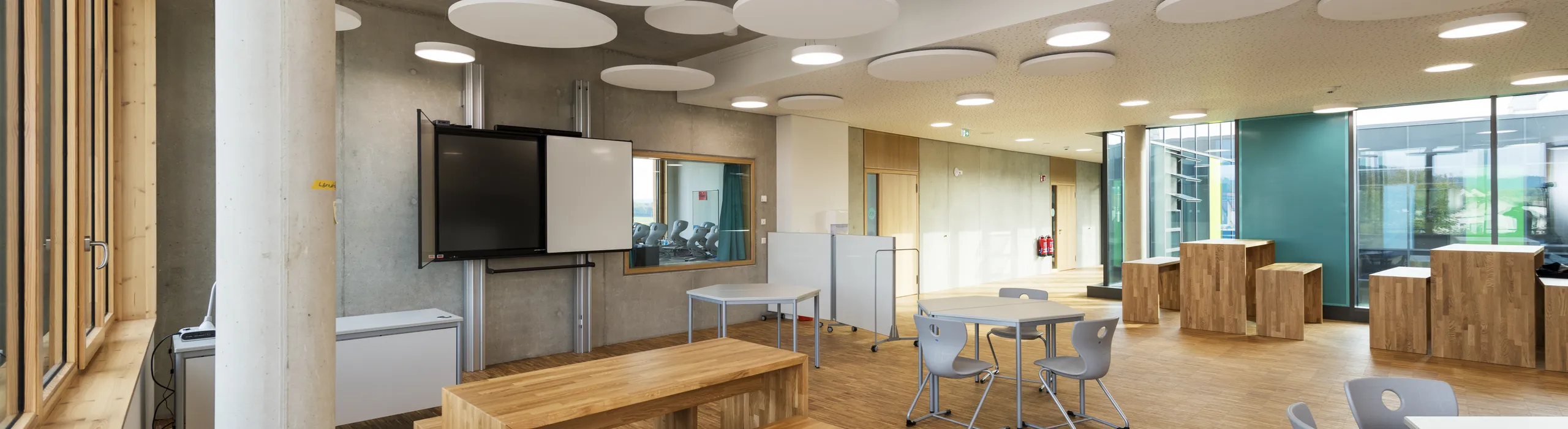 Modern classroom with wooden floors, benches, circular ceiling panels, a large screen, and blue-green walls. Natural light from large windows.