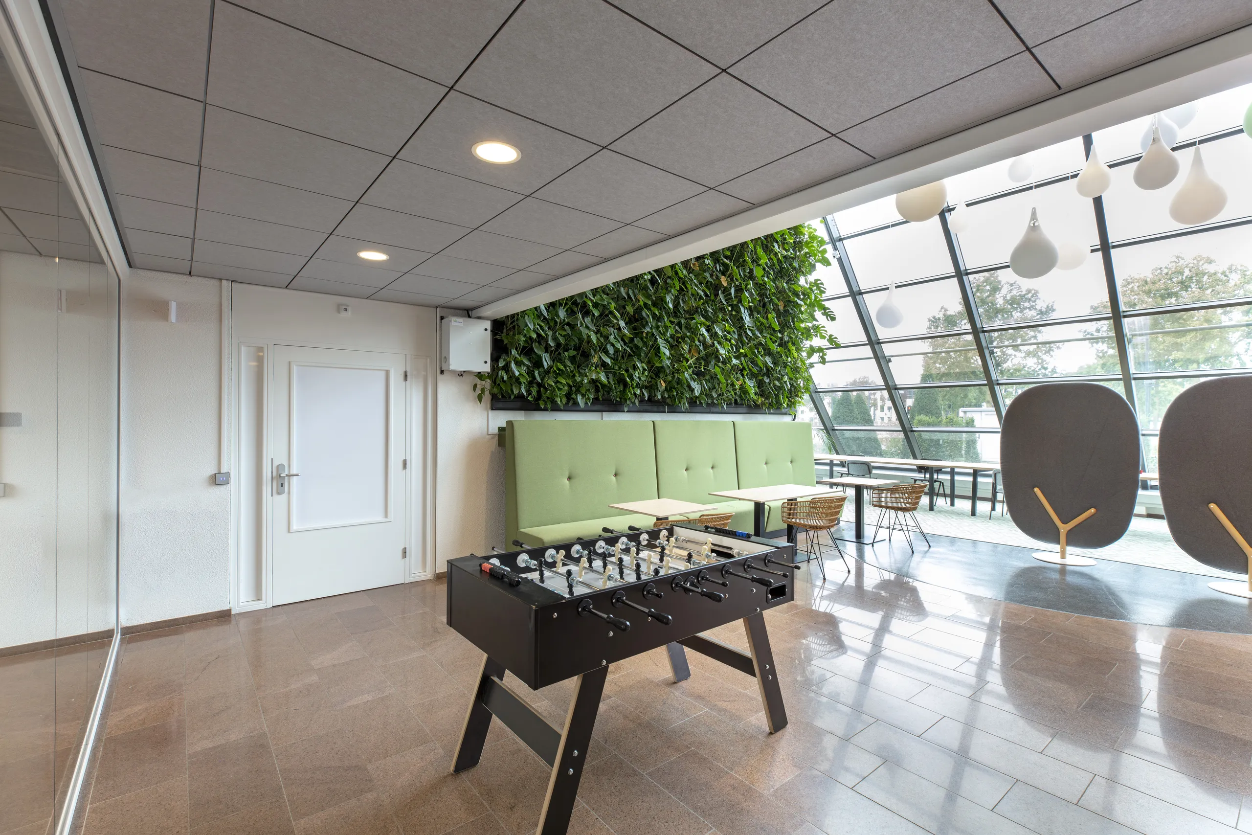 Office breakout area with grey acoustic ceiling tiles, foosball table, green seating, a living plant wall, and large windows bringing in natural daylight.