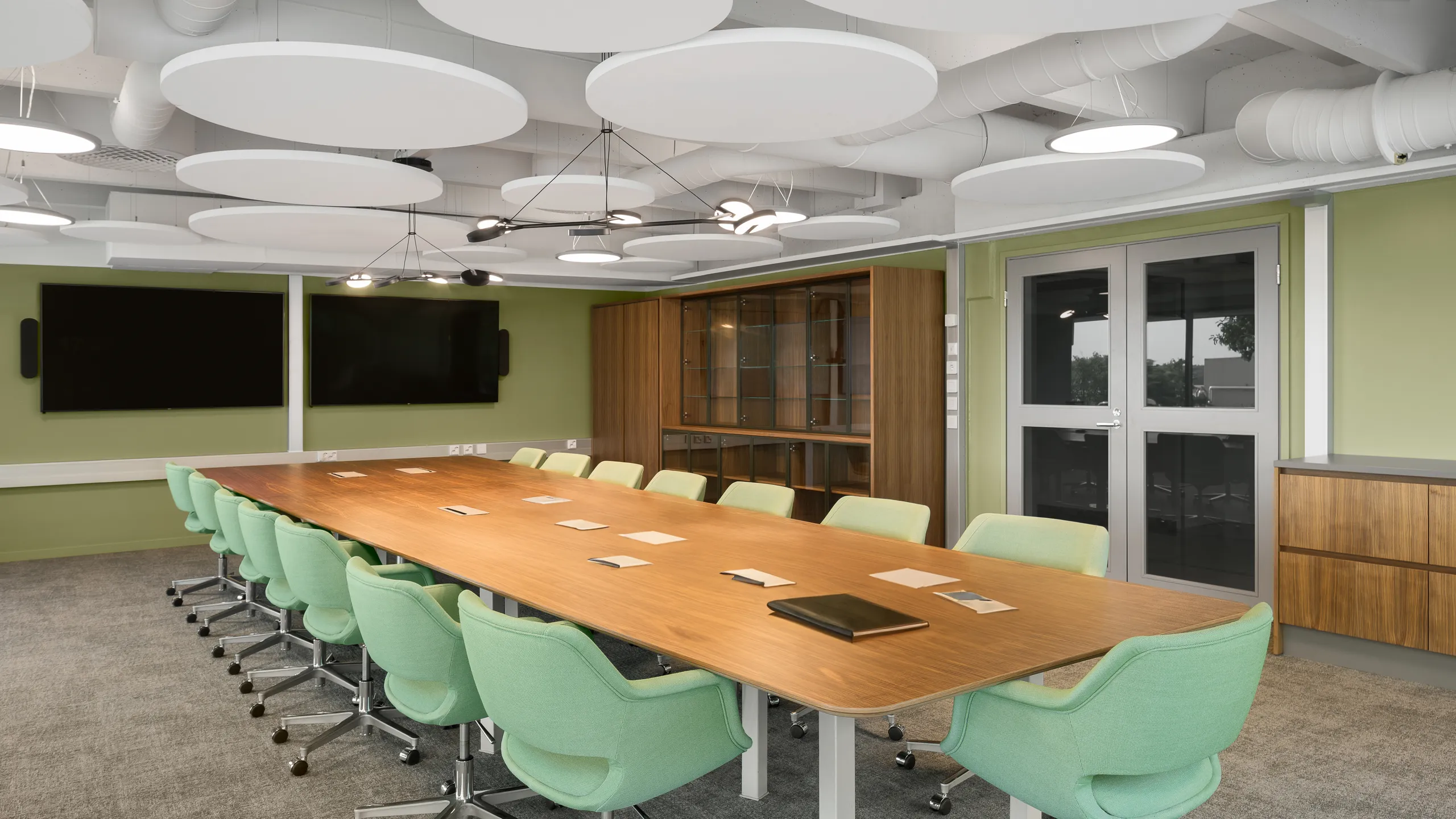 Meeting room with suspended circular acoustic rafts, large conference table, green chairs, and integrated screens in a modern office setting.