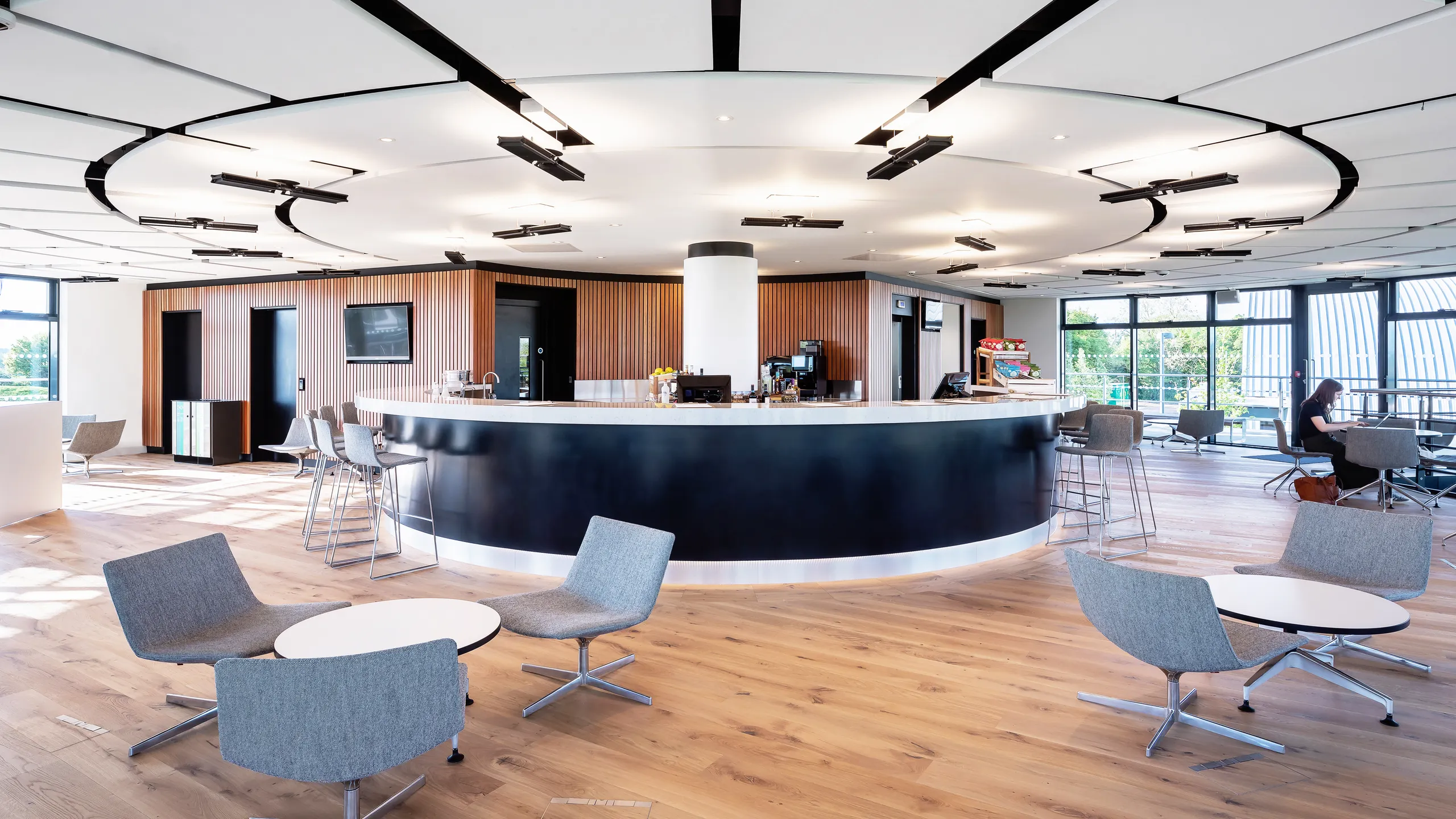 Modern open-concept lounge with floating raft ceilings, central circular counter, surrounded by gray chairs and small tables on a wooden floor, featuring large windows.