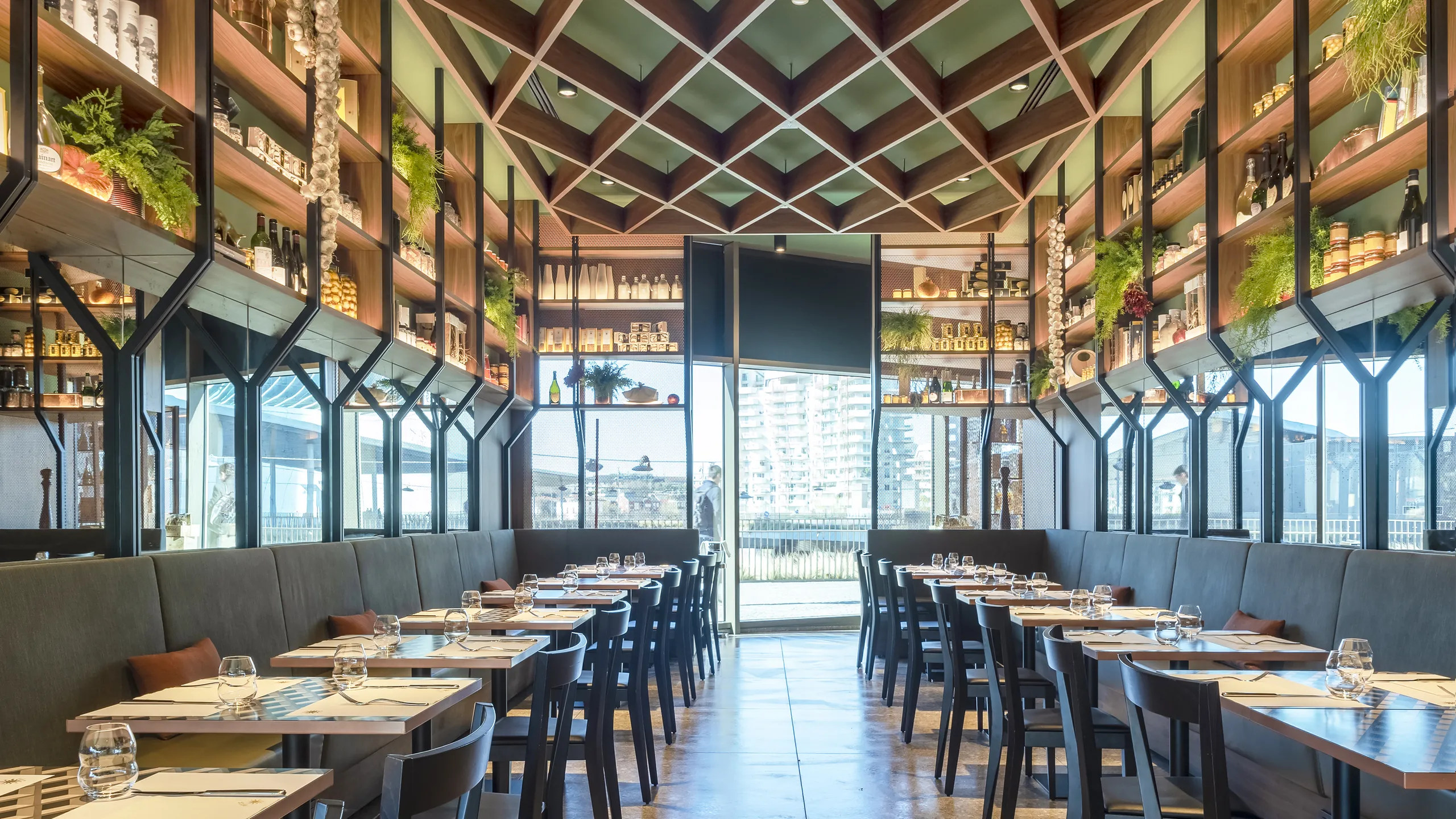 Modern restaurant interior with wooden shelves, green ceiling, and neatly arranged tables. Large window at the back offers a view outside.