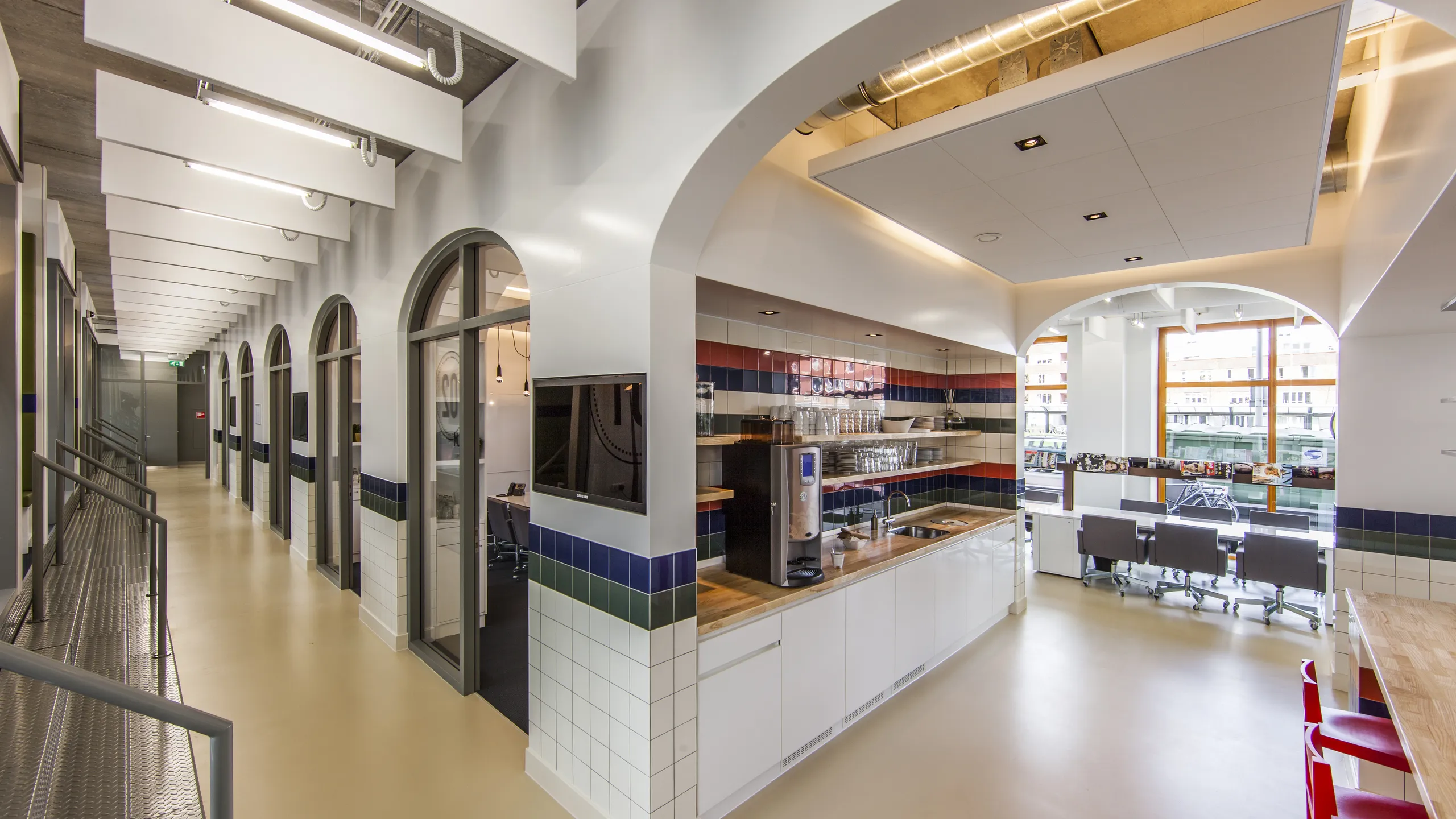 Office corridor and kitchen area with arched openings, white acoustic ceiling baffles along the hallway, and a suspended acoustic ceiling island above the kitchenette.