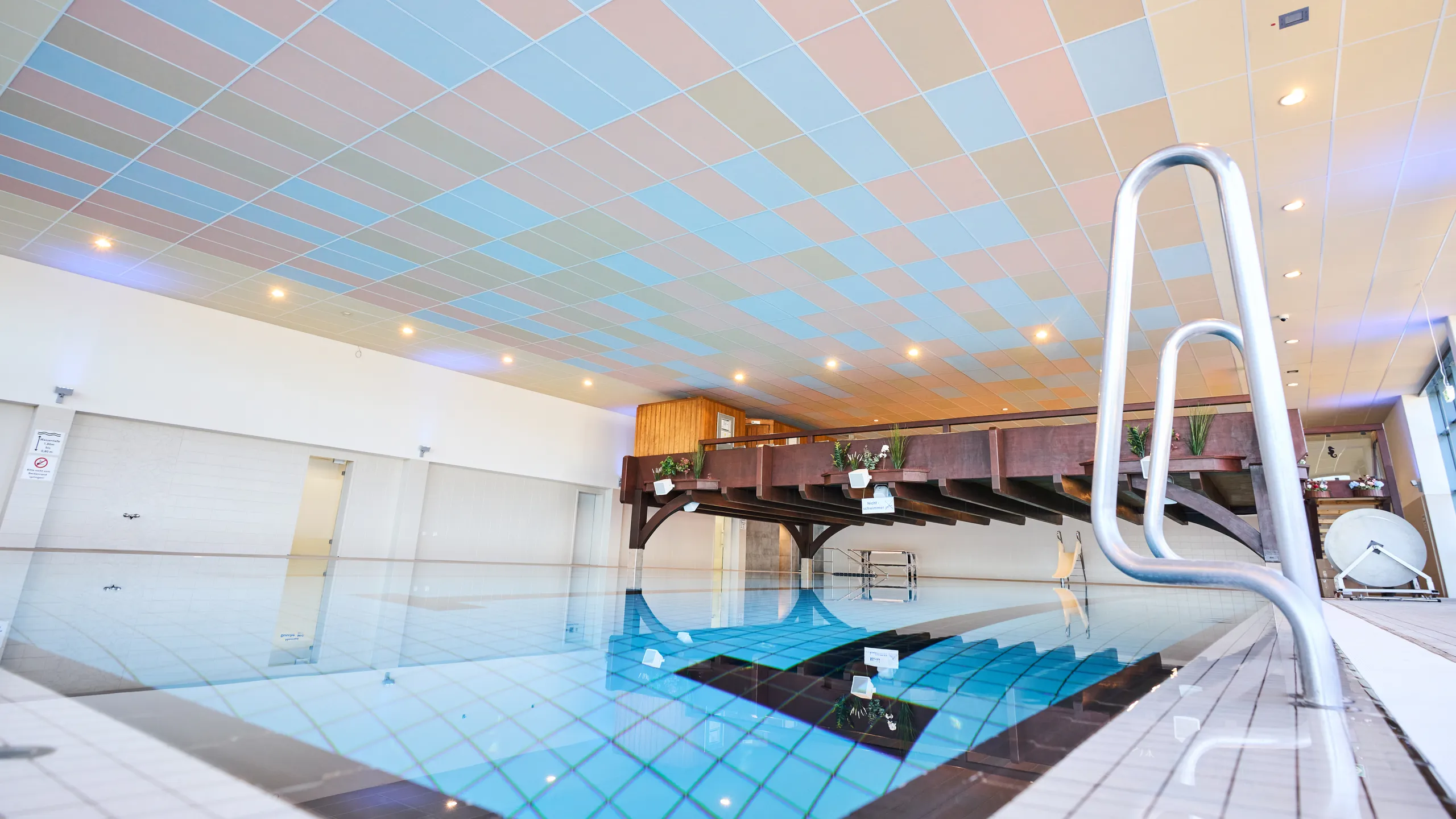 Indoor swimming pool with a pastel-coloured acoustic tile ceiling, calm water surface, and a wooden poolside deck overlooking the pool area.