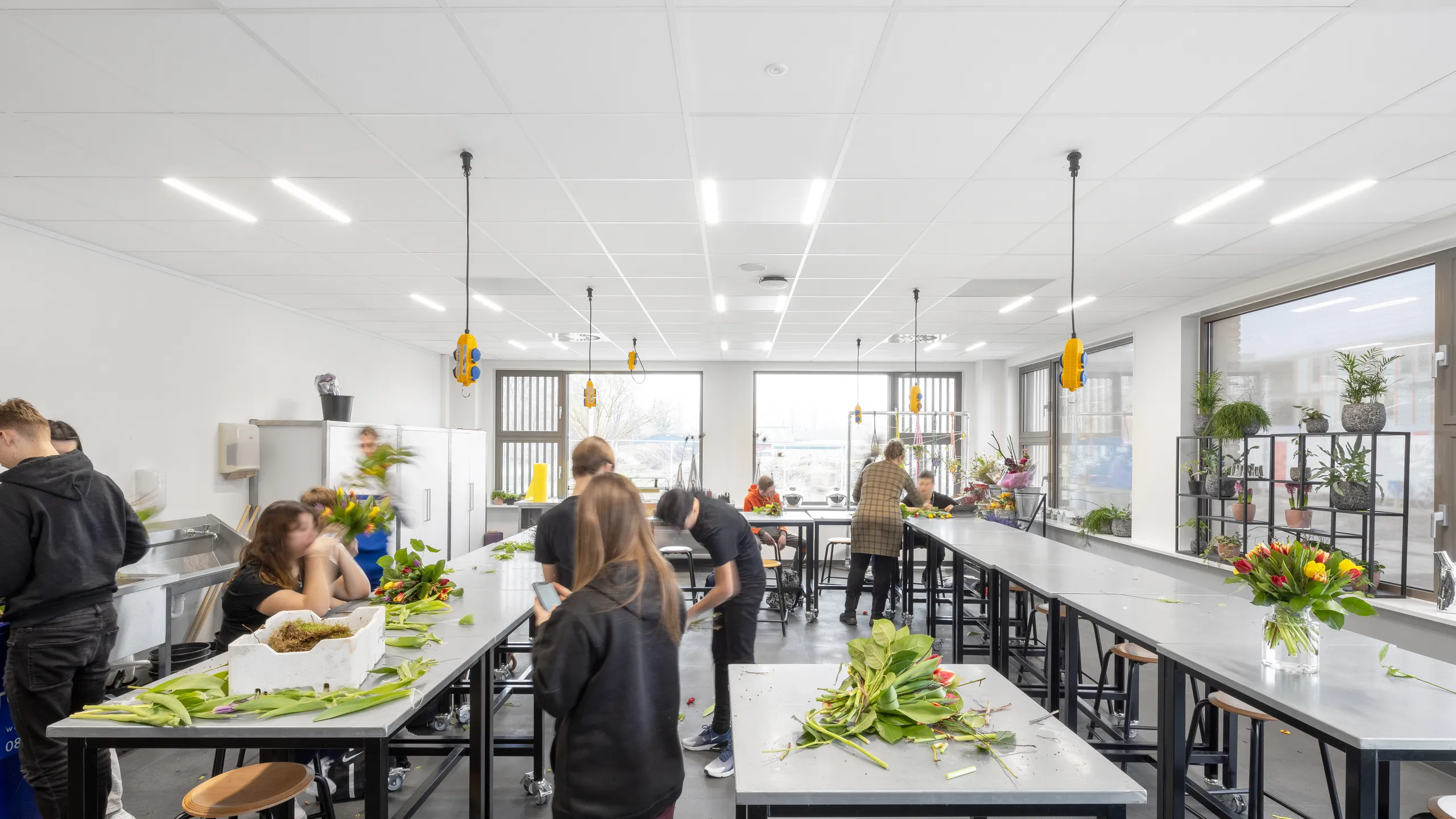People arranging flowers in a bright, spacious room with white tabletops and large windows. Plants and bouquets adorn the space.