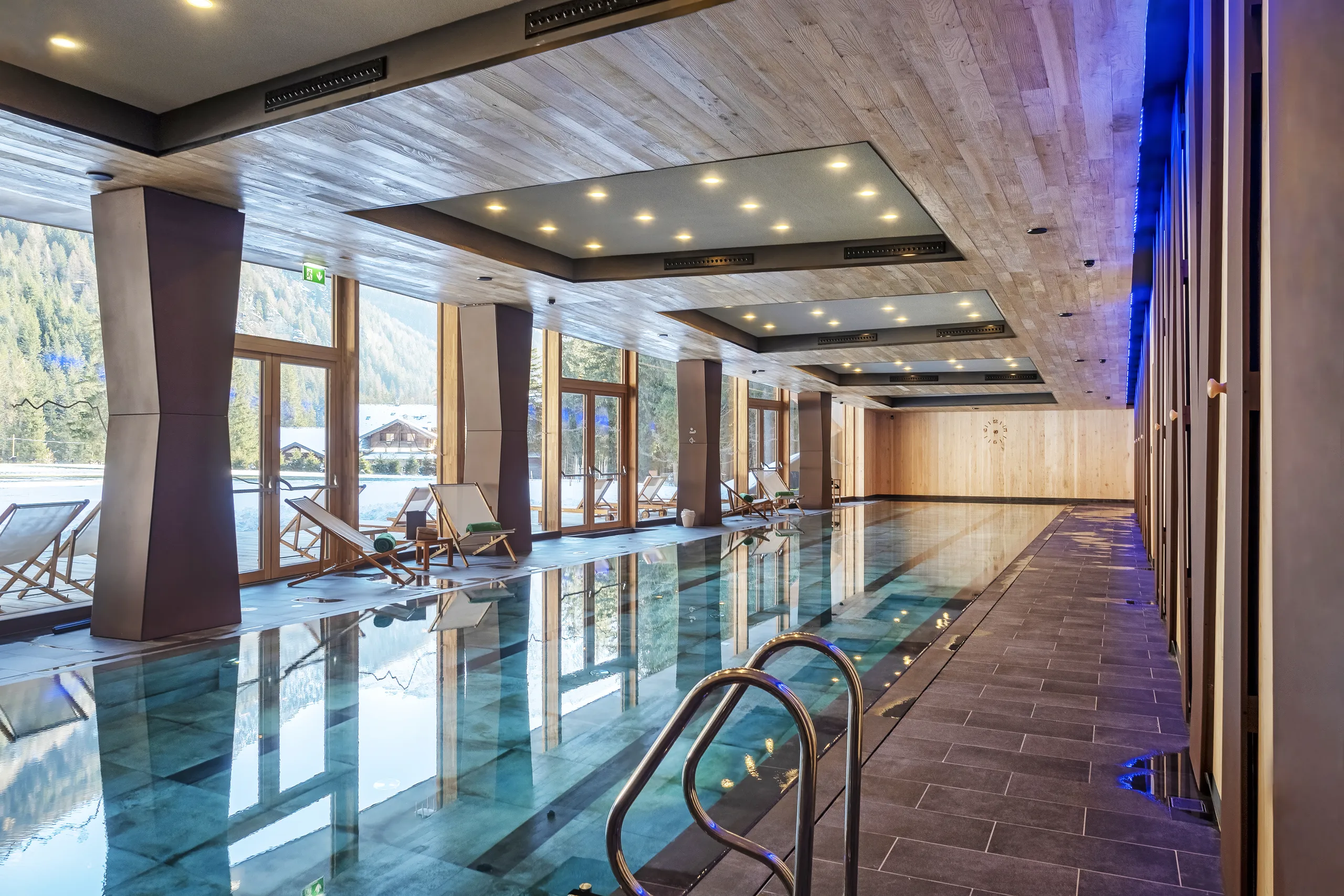 Indoor swimming pool with large floor-to-ceiling windows, wooden ceiling panels with integrated lighting, and lounge chairs overlooking a snowy mountain landscape.