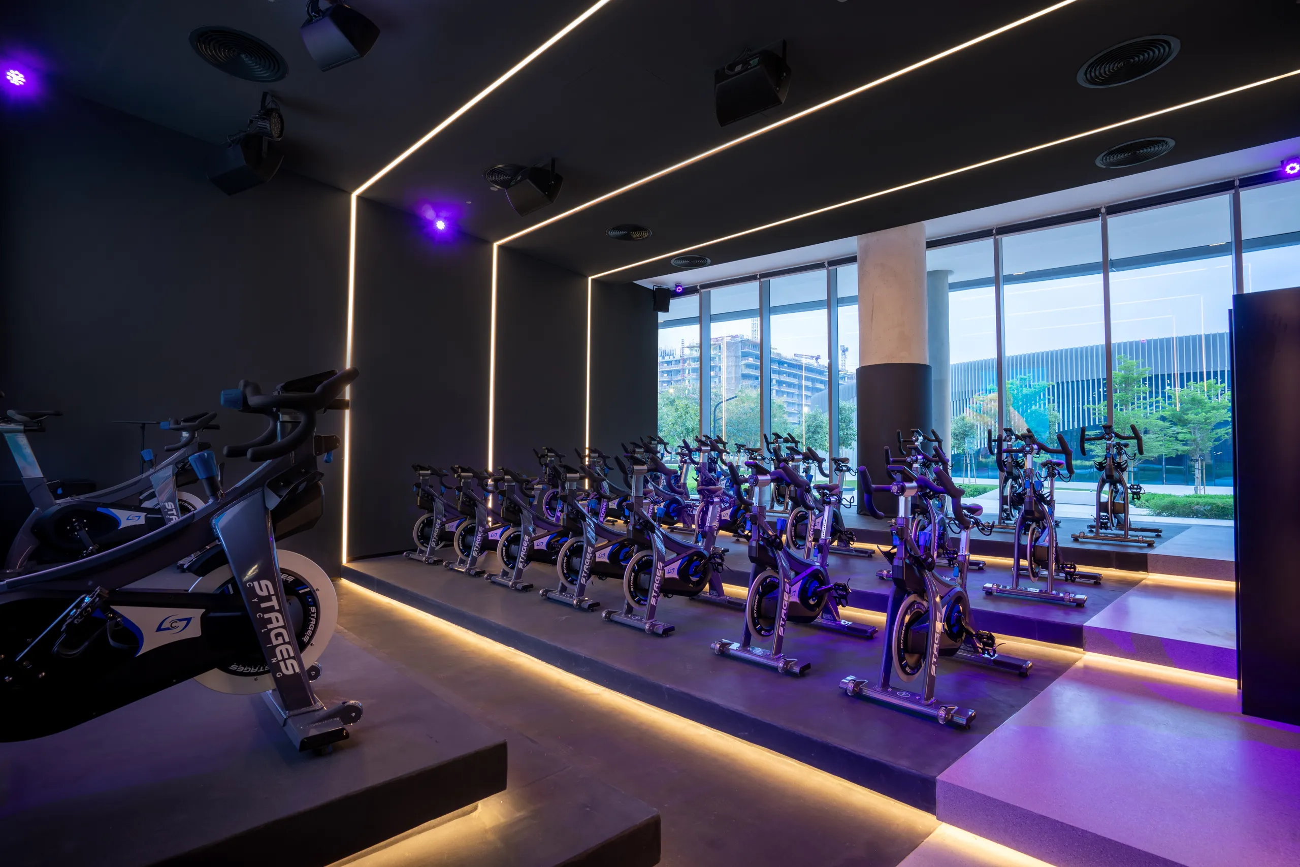 Indoor cycling studio with rows of stationary bikes on a raised platform, LED strip lighting on dark walls and ceiling, and large windows overlooking an outdoor urban landscape.