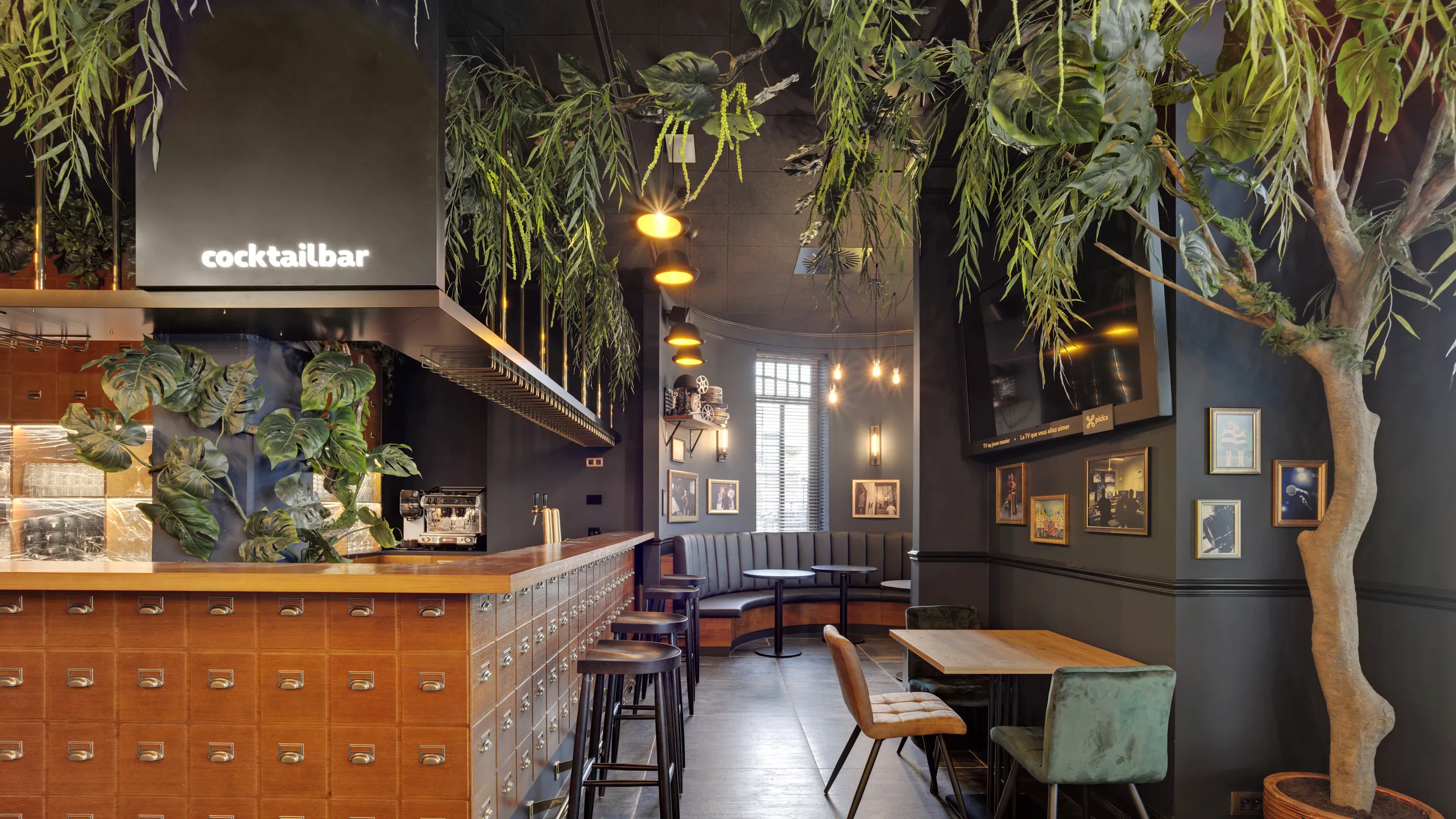 Stylish cocktail bar with black suspended ceiling, wooden counter, high stools, and lush greenery. Cozy seating area with upholstered chairs and framed art on walls.